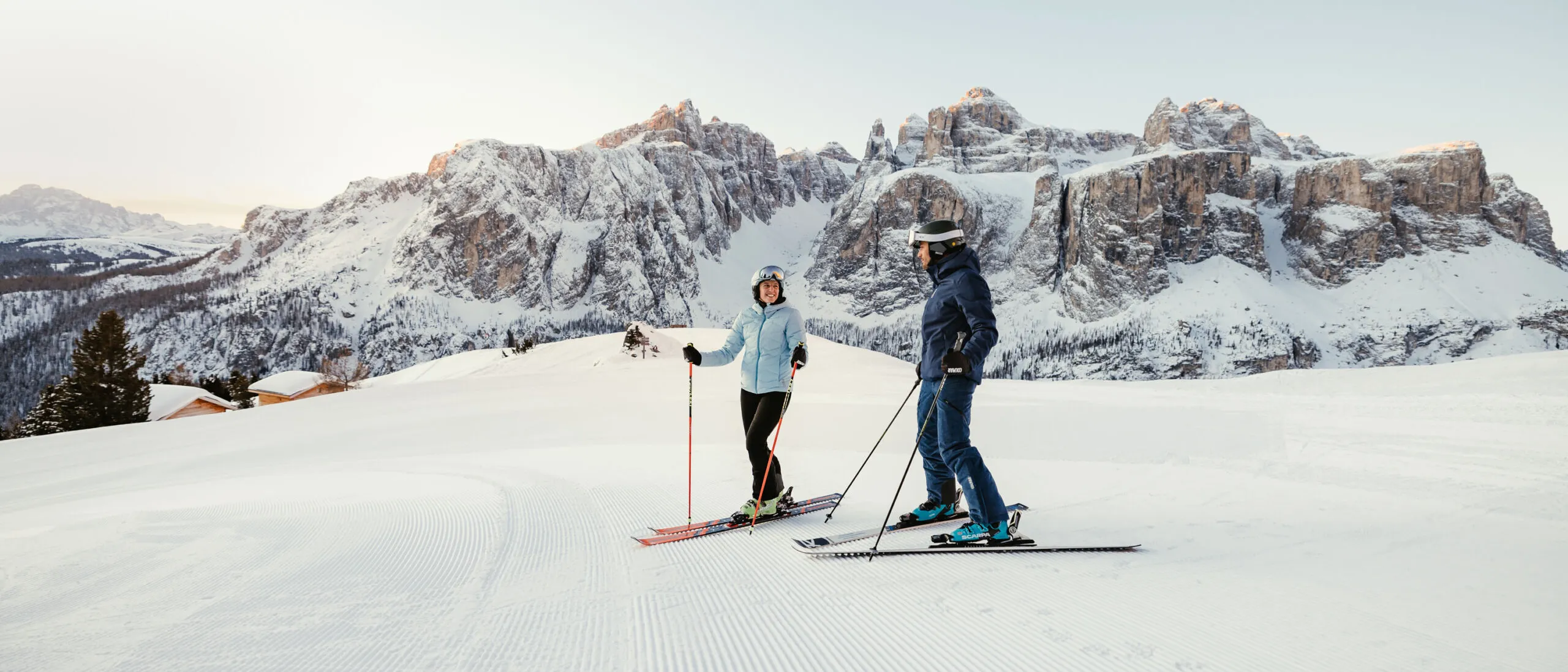 Two skiers in light blue and dark blue jackets stand on a groomed snowy slope with mountainous backdrop.