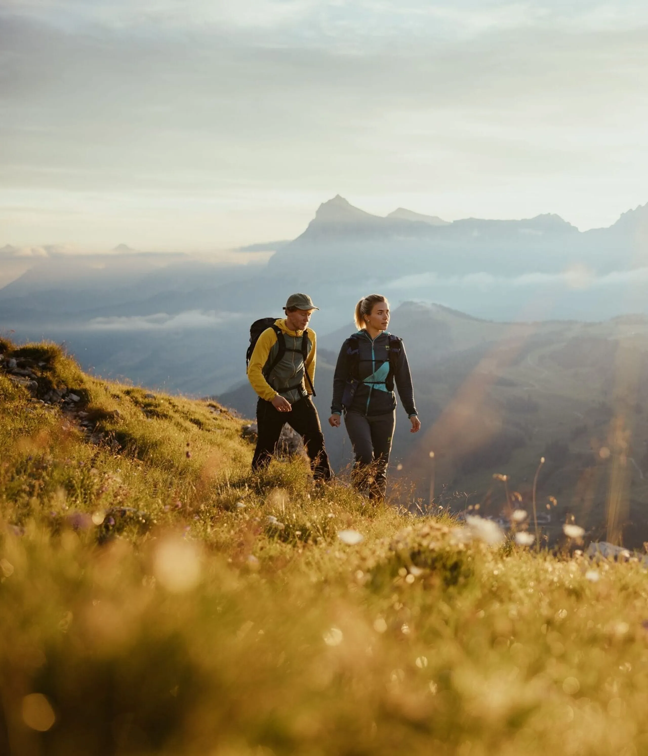Two hikers trek along a sunlit alpine meadow with mountains in the distance during golden hour.