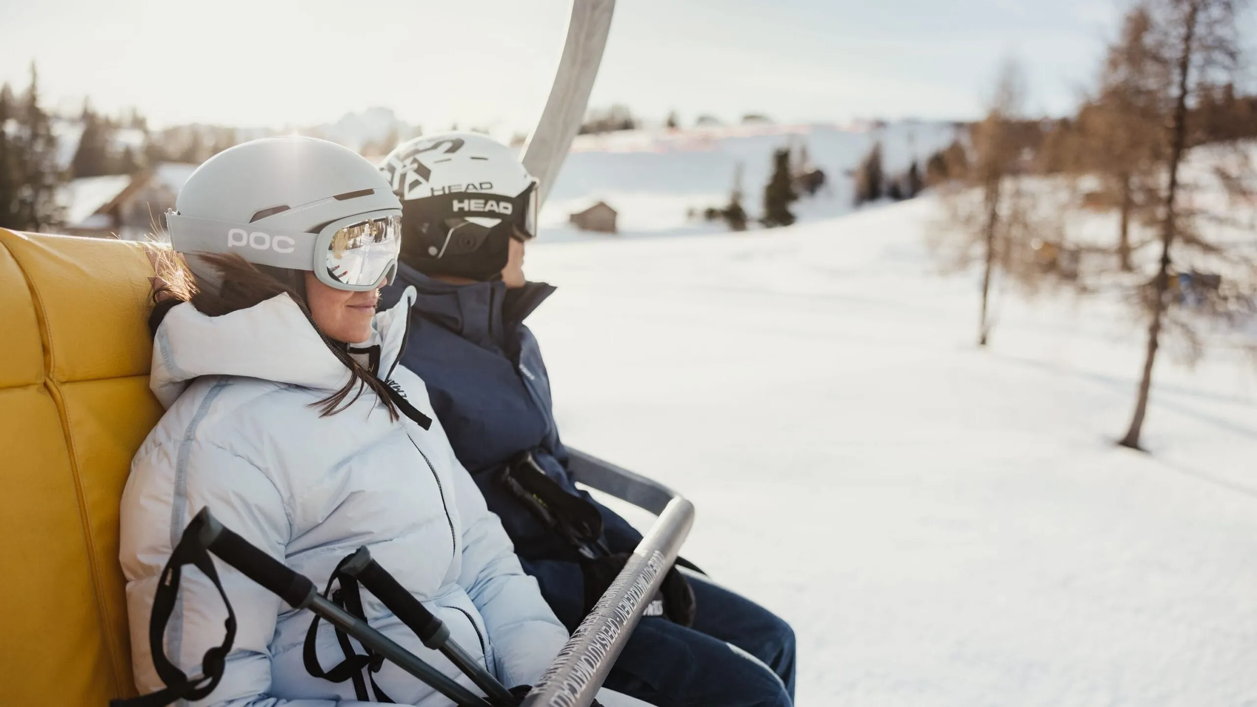Two people in ski gear sit on a yellow slope chairlift against a snowy alpine landscape with trees in the background.