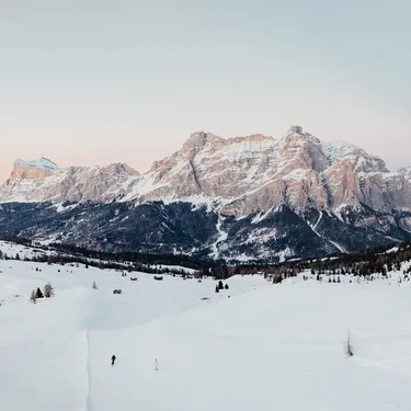 Snowy alpine valley with distant rugged mountains under pale sky at sunset, scattered trees and lone skiers.