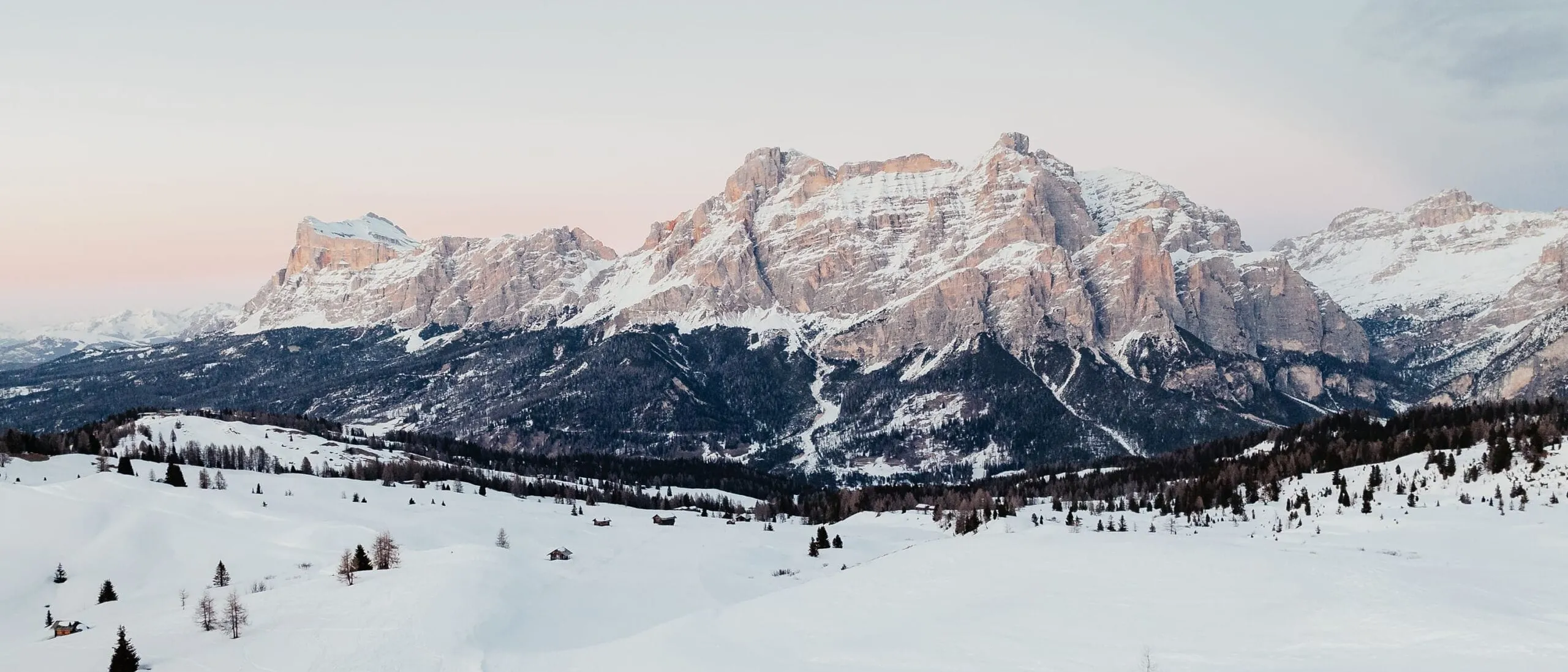 Snowy alpine valley with distant rugged mountains under pale sky at sunset, scattered trees and lone skiers.