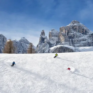 Skiers descend snowy slope with towering rocky alpine peaks under blue sky.