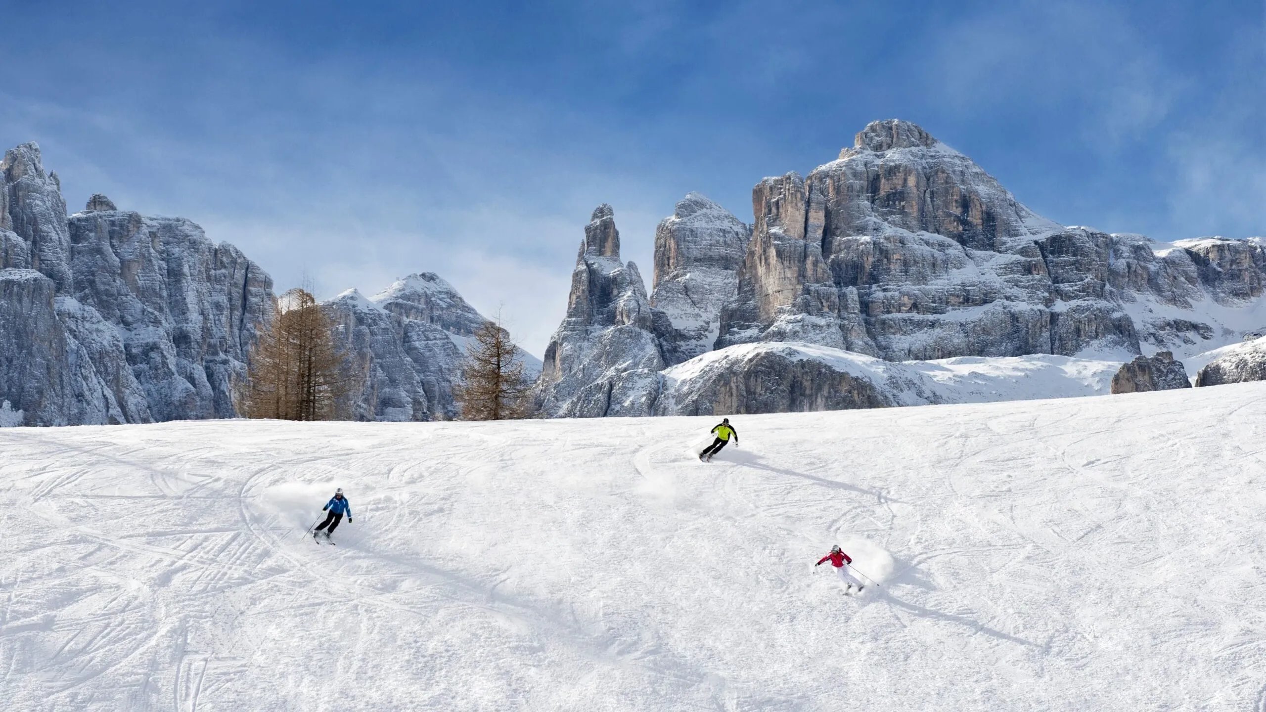 Skiers descend snowy slope with towering rocky alpine peaks under blue sky.