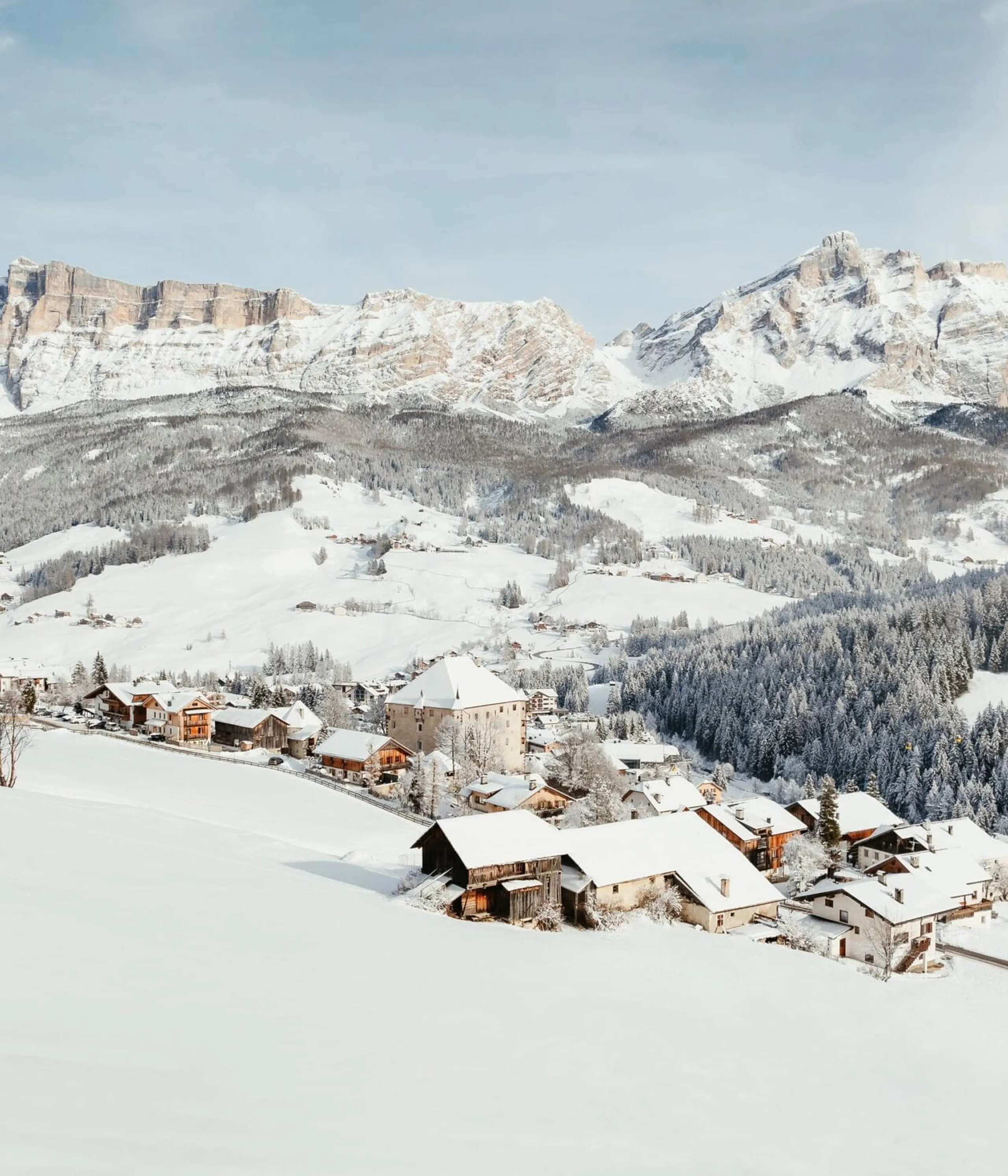 Snow-covered alpine village nestled in a valley with church, chalets, and forested slopes beneath towering snowy peaks.