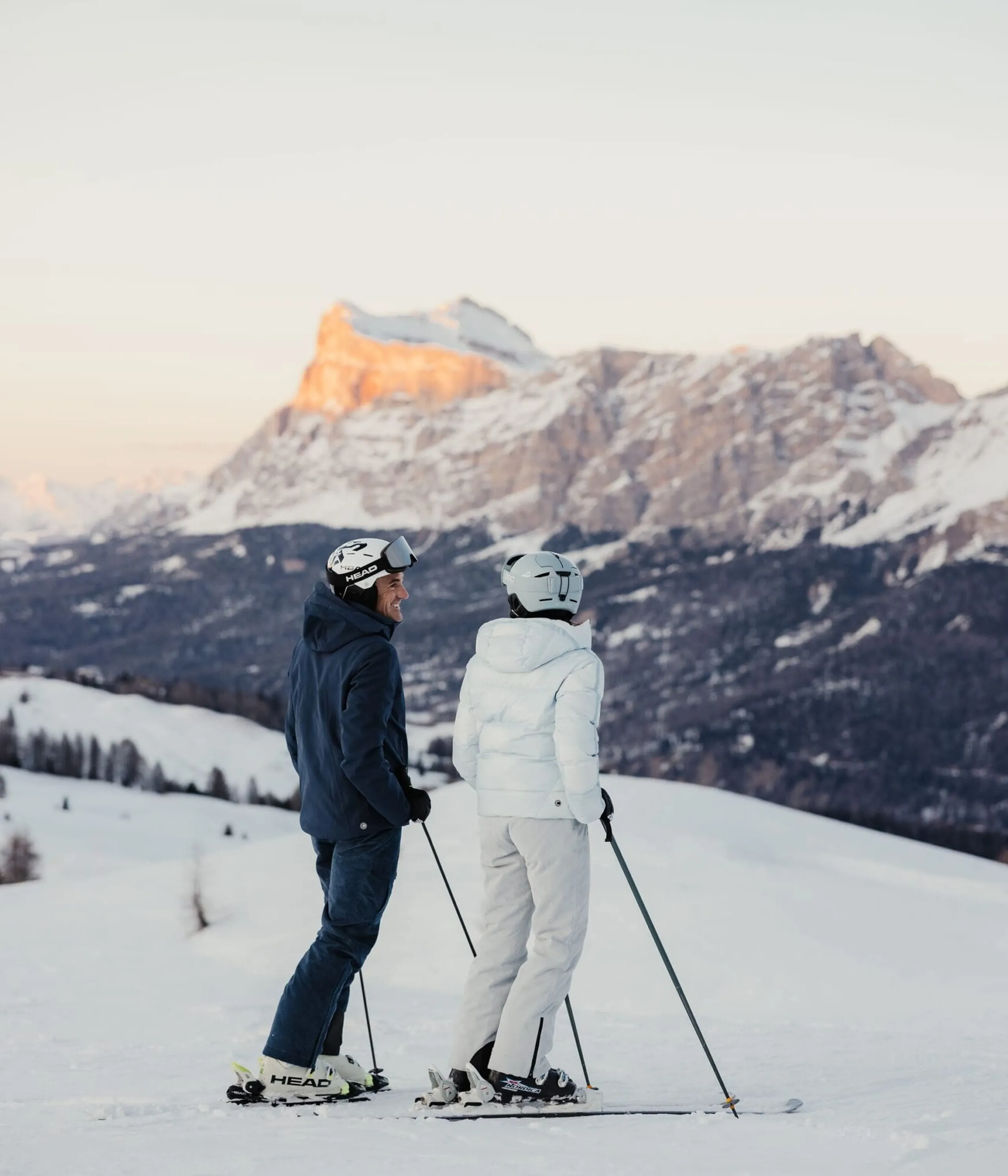 Two skiers in dark and white gear stand with poles on snowy slope, facing snowy mountains at sunset.