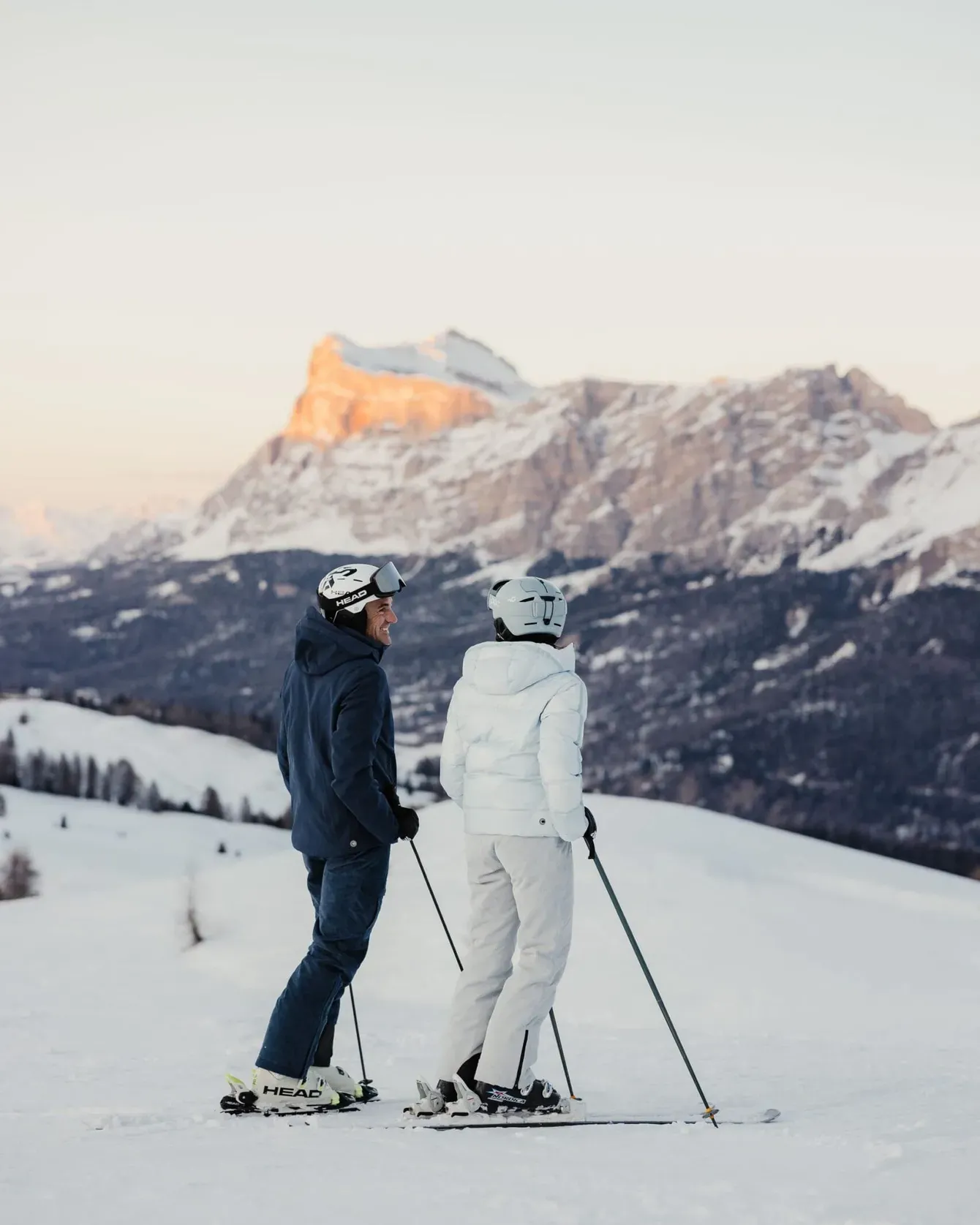 Two skiers in dark and white gear stand with poles on snowy slope, facing snowy mountains at sunset.