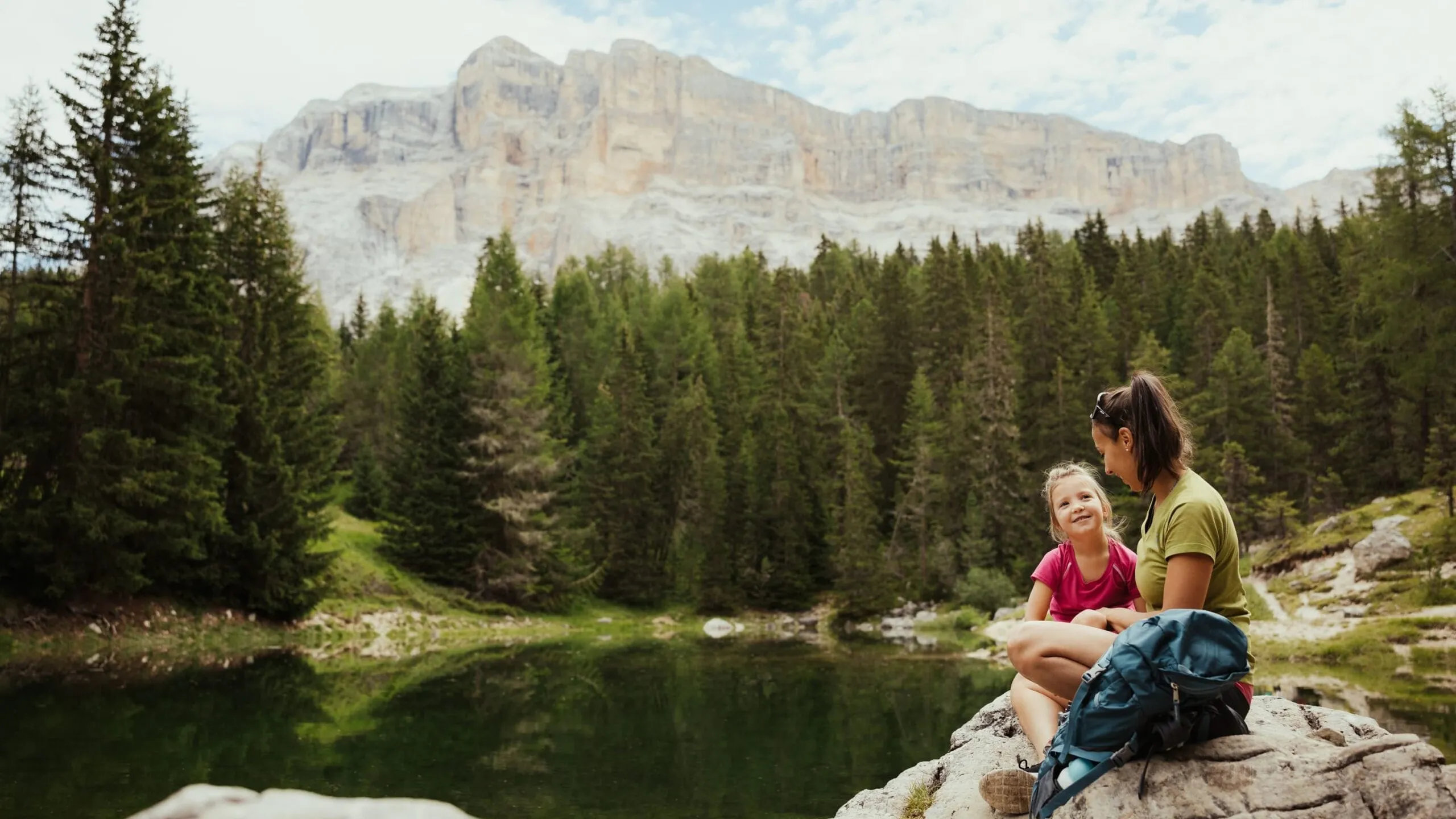 Two hikers, a woman and a child, sit on rocks by a calm alpine lake with forested mountains in the background.