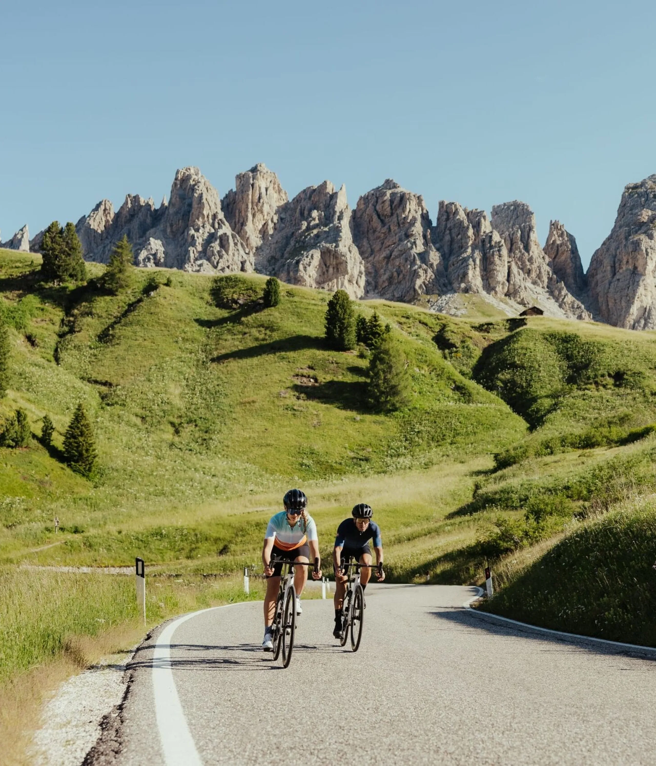 Two cyclists ride a winding road through grassy hills with jagged rocky mountains in the background under a clear blue sky.