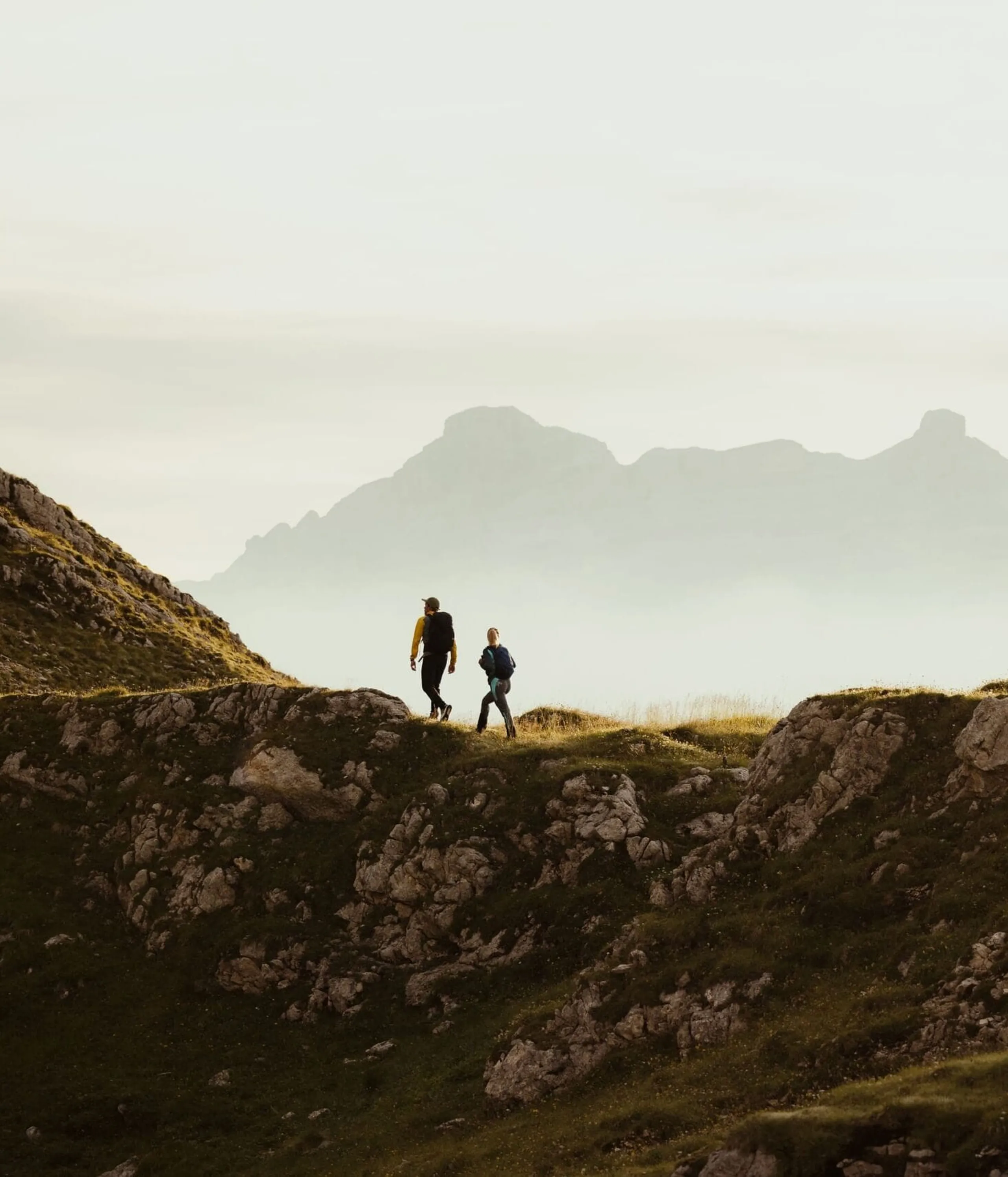 Two hikers walk along a rocky ridge with distant misty mountains in the background.