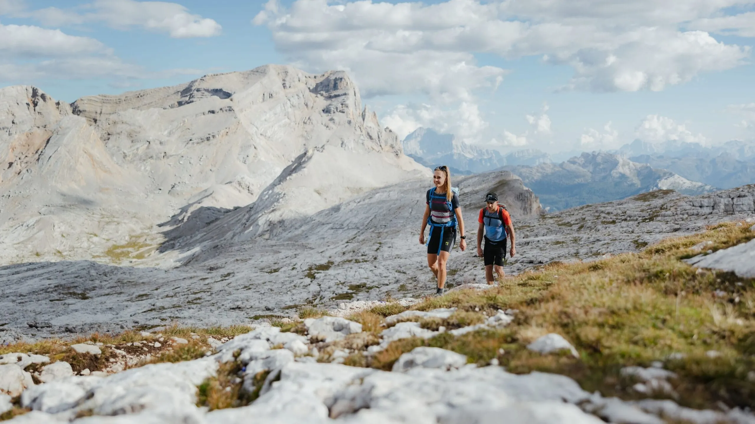 Hikers trek along a rocky alpine path with towering gray mountains and blue skies in the background.