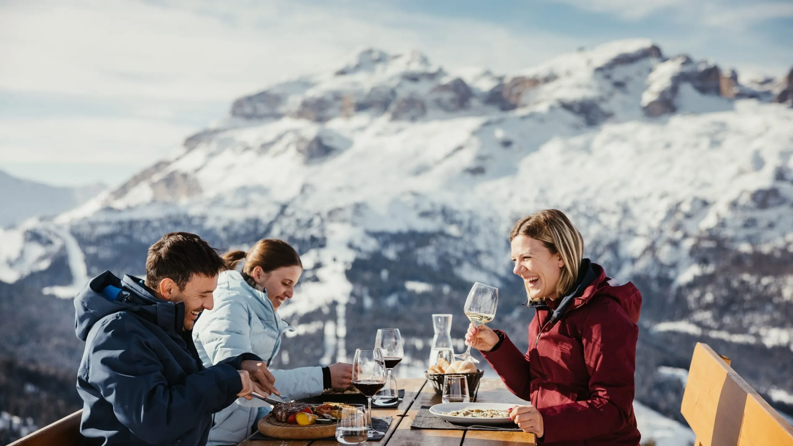 Outdoor alpine meal with three people enjoying wine at a wooden table against snowy mountains.