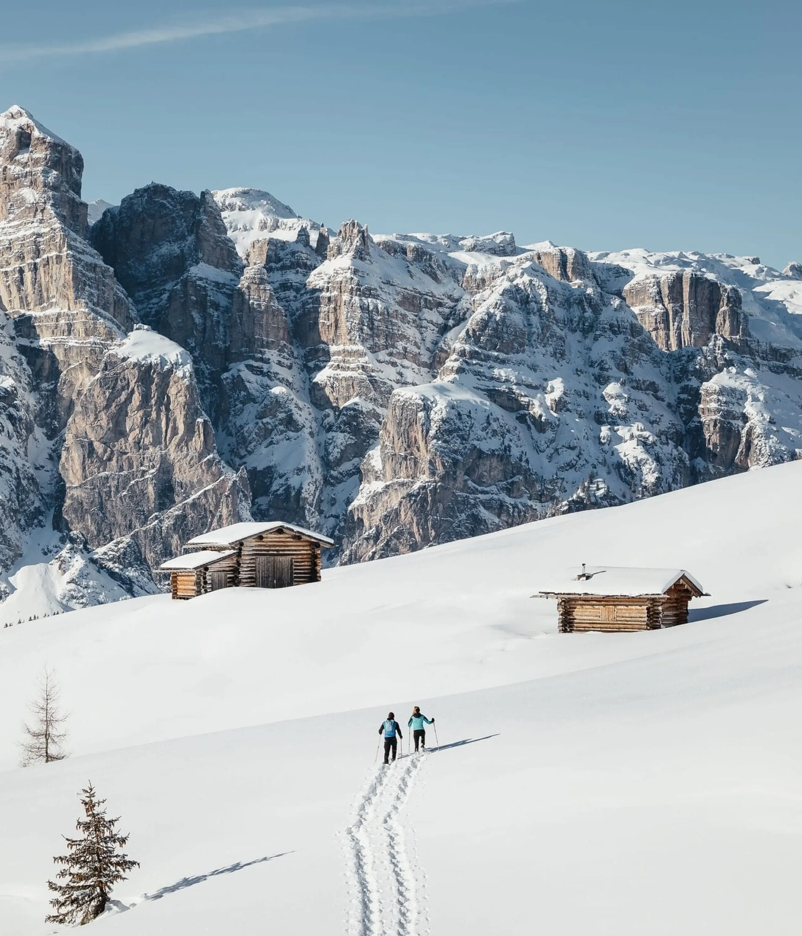 Two skiers trek across a snowy mountainside toward wooden cabins with towering rocky peaks in the background.