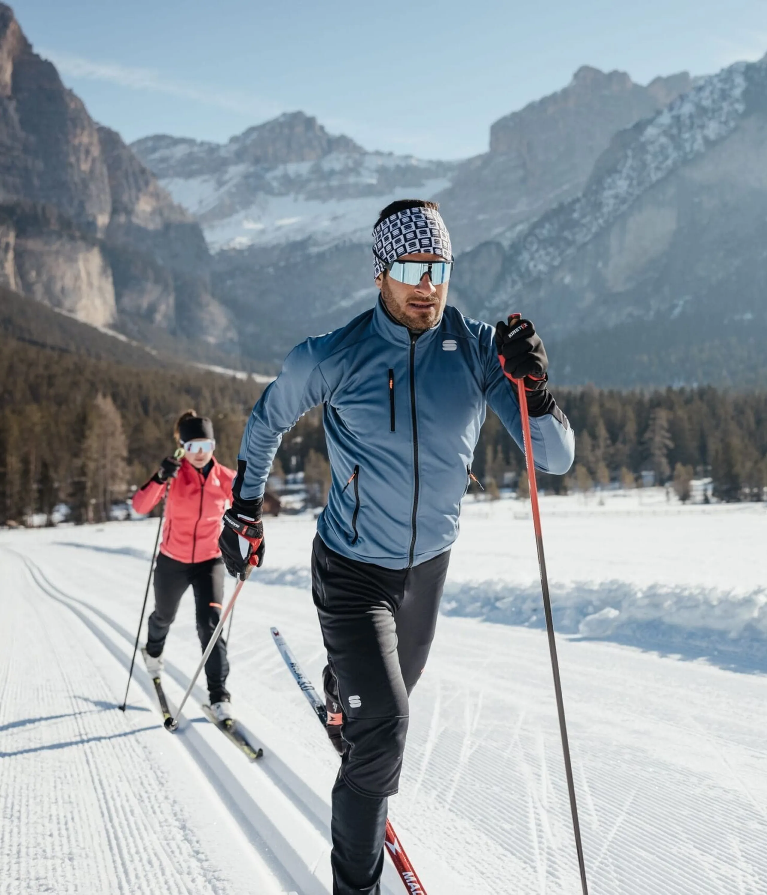 Cross-country skiers glide on a snowy trail with mountains and pine trees in the background.