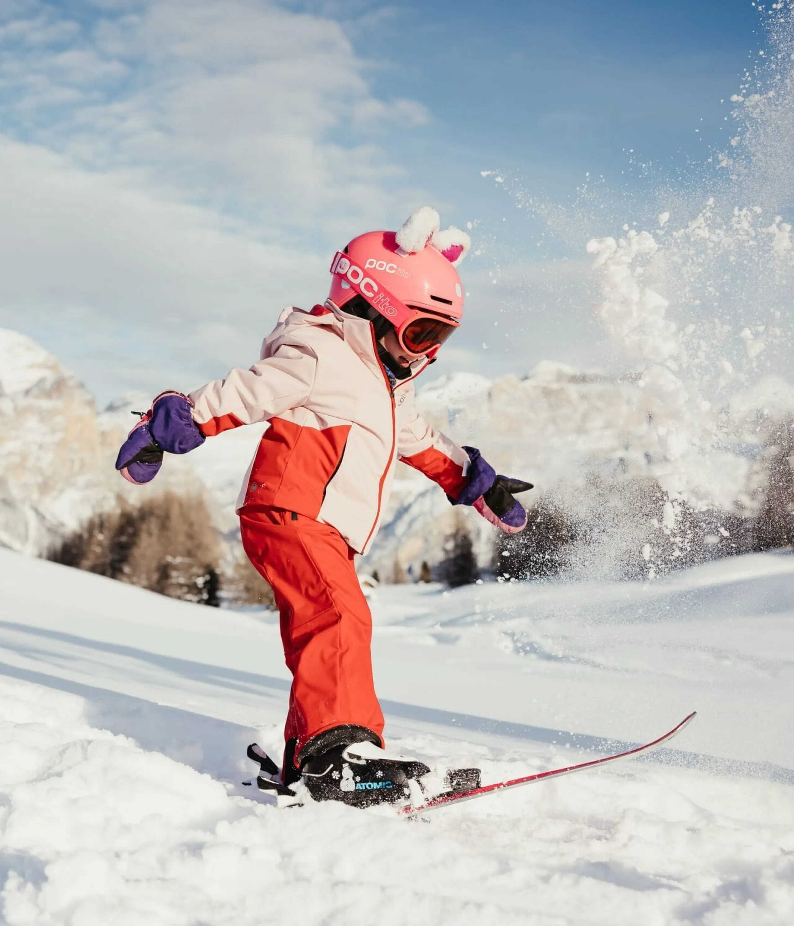 Child wearing pink helmet and red snowsuit skis down snowy slope, kicking up powder in alpine landscape.