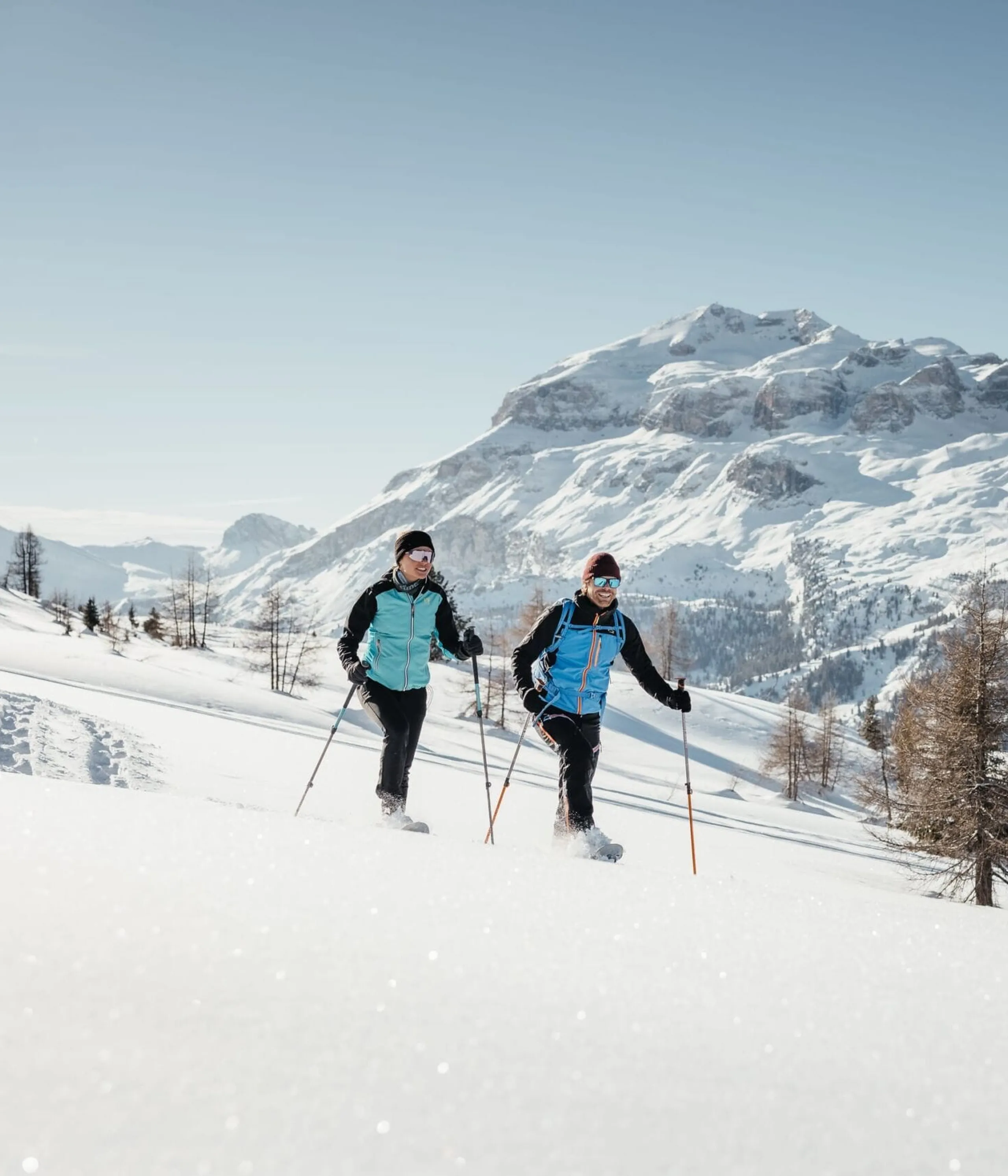 Two hikers in turquoise jackets ski touring across a snowy alpine landscape with rugged mountains in the background.