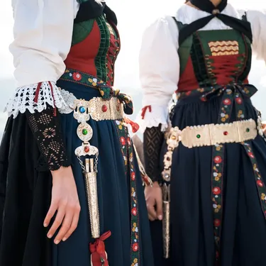 Two people in traditional Alpine folk costumes with embroidered bodices, lace cuffs, and jeweled belts standing by a wooden fence.