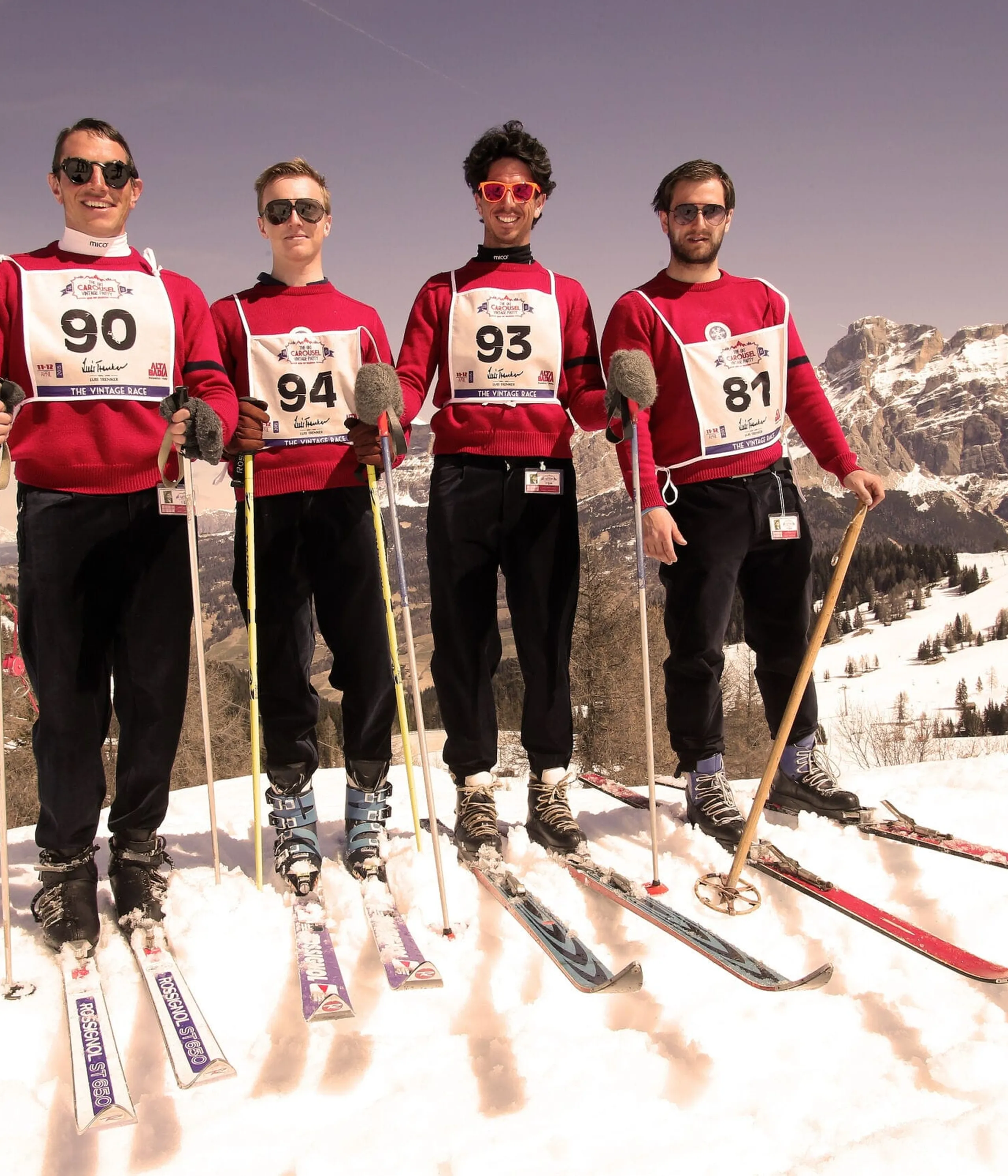 Four skiers in red tops with race bibs standing on snowy slope with mountains in the background.