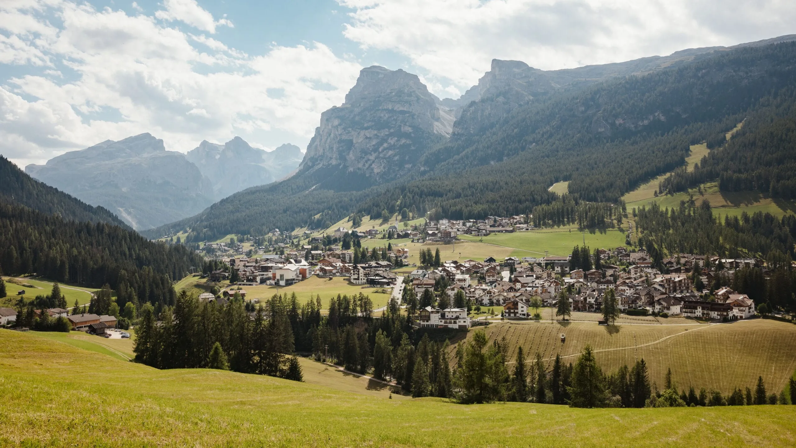 A mountain valley town with scattered houses, green fields, and dense forests set against rugged cliffs under a partly cloudy sky.