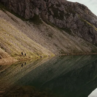 Two hikers stand on a rocky lakeshore, mirrored by the calm mountain lake and steep cliff behind them.