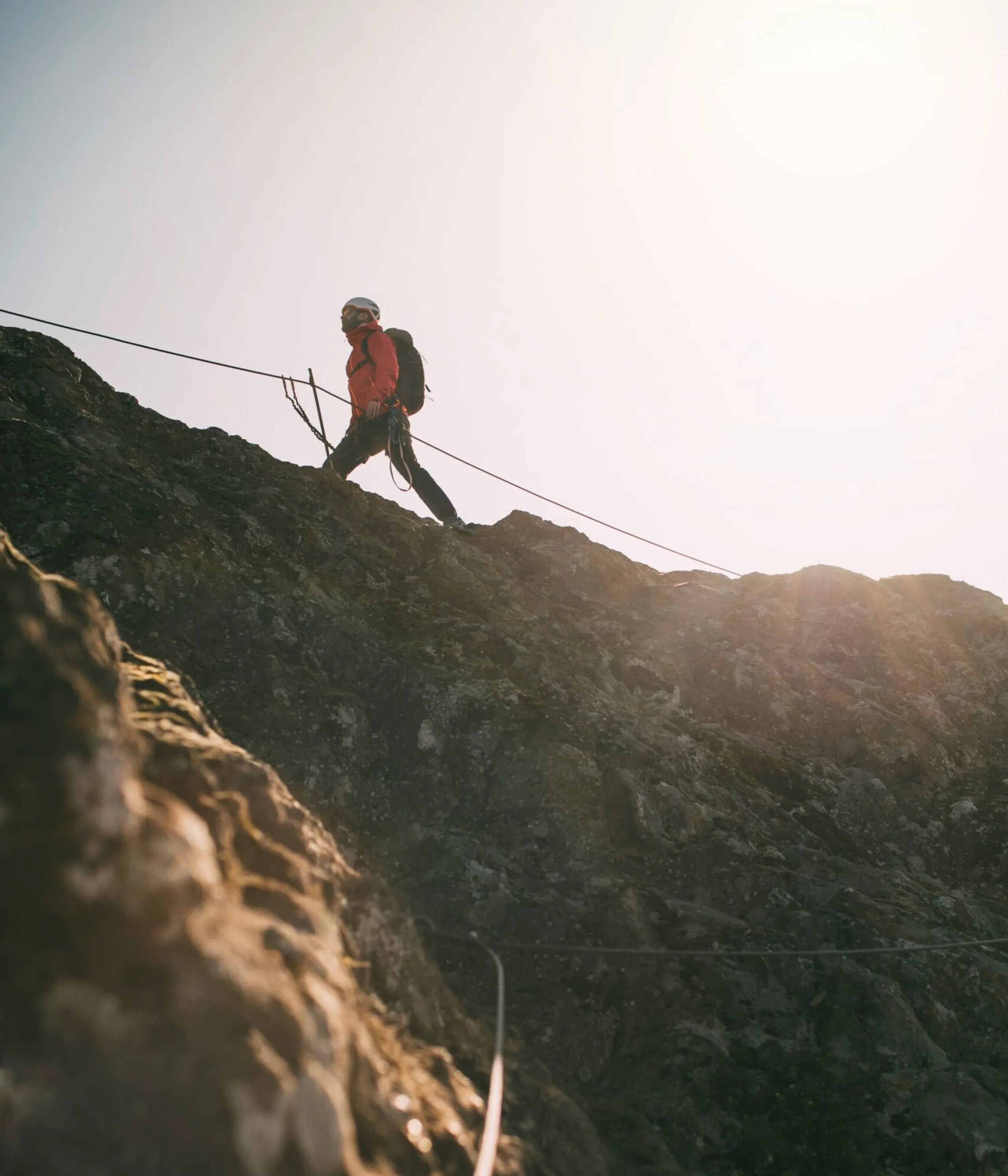 Rocky mountain climber in red jacket ascending a steep cliff with safety ropes at sunrise.