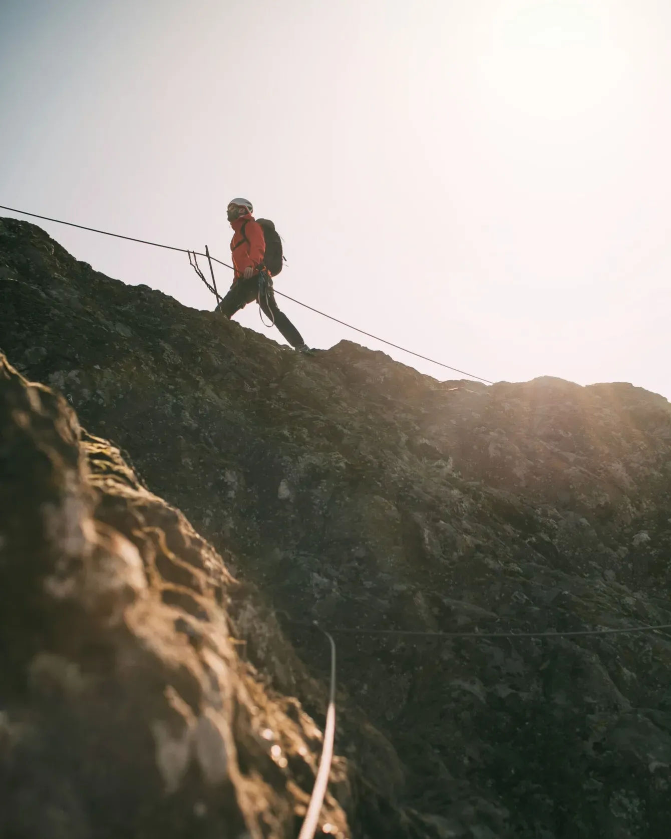Rocky mountain climber in red jacket ascending a steep cliff with safety ropes at sunrise.