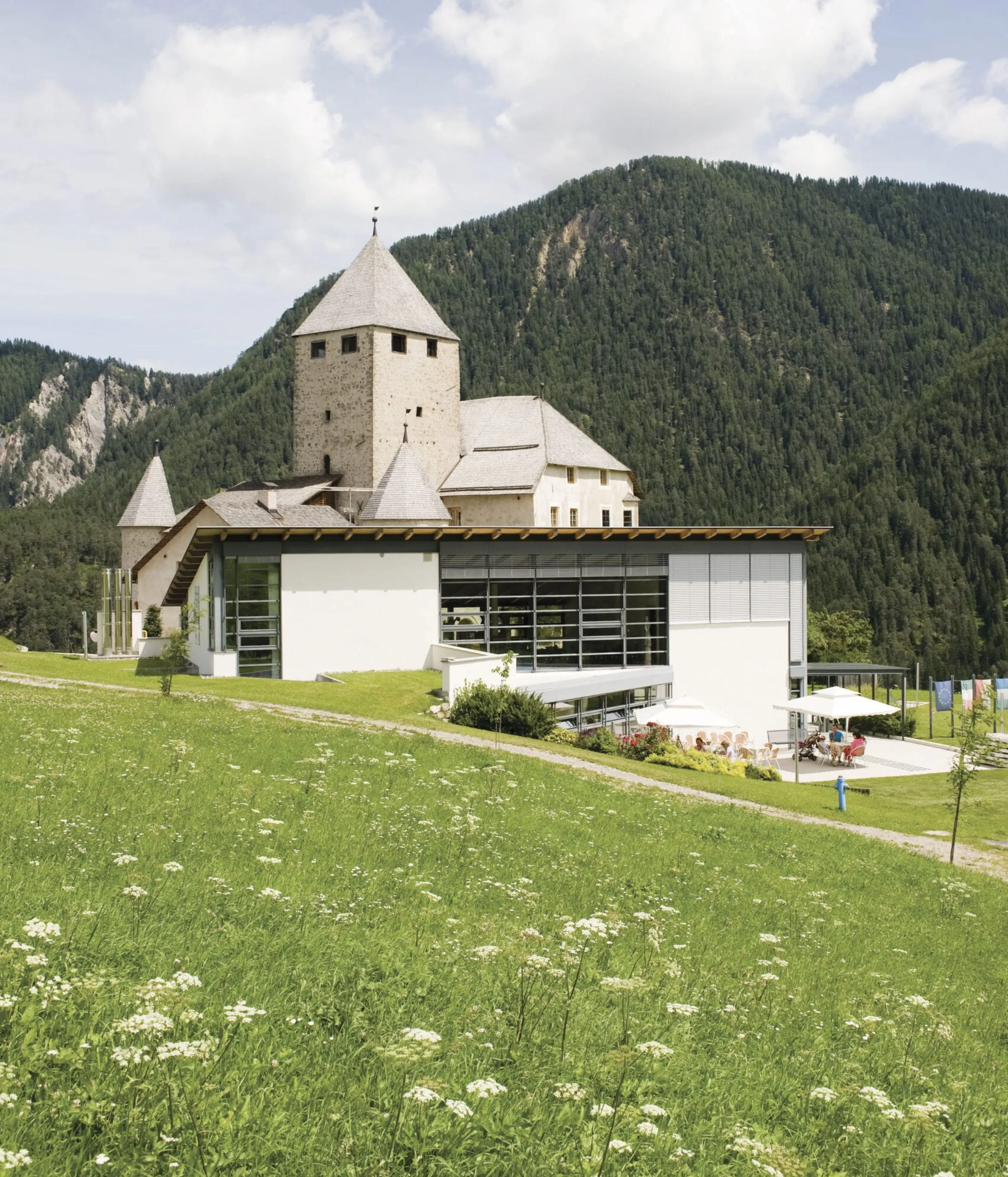 Castle-style stone building with conical towers sits on a grassy meadow against forested hills and a modern glass extension.