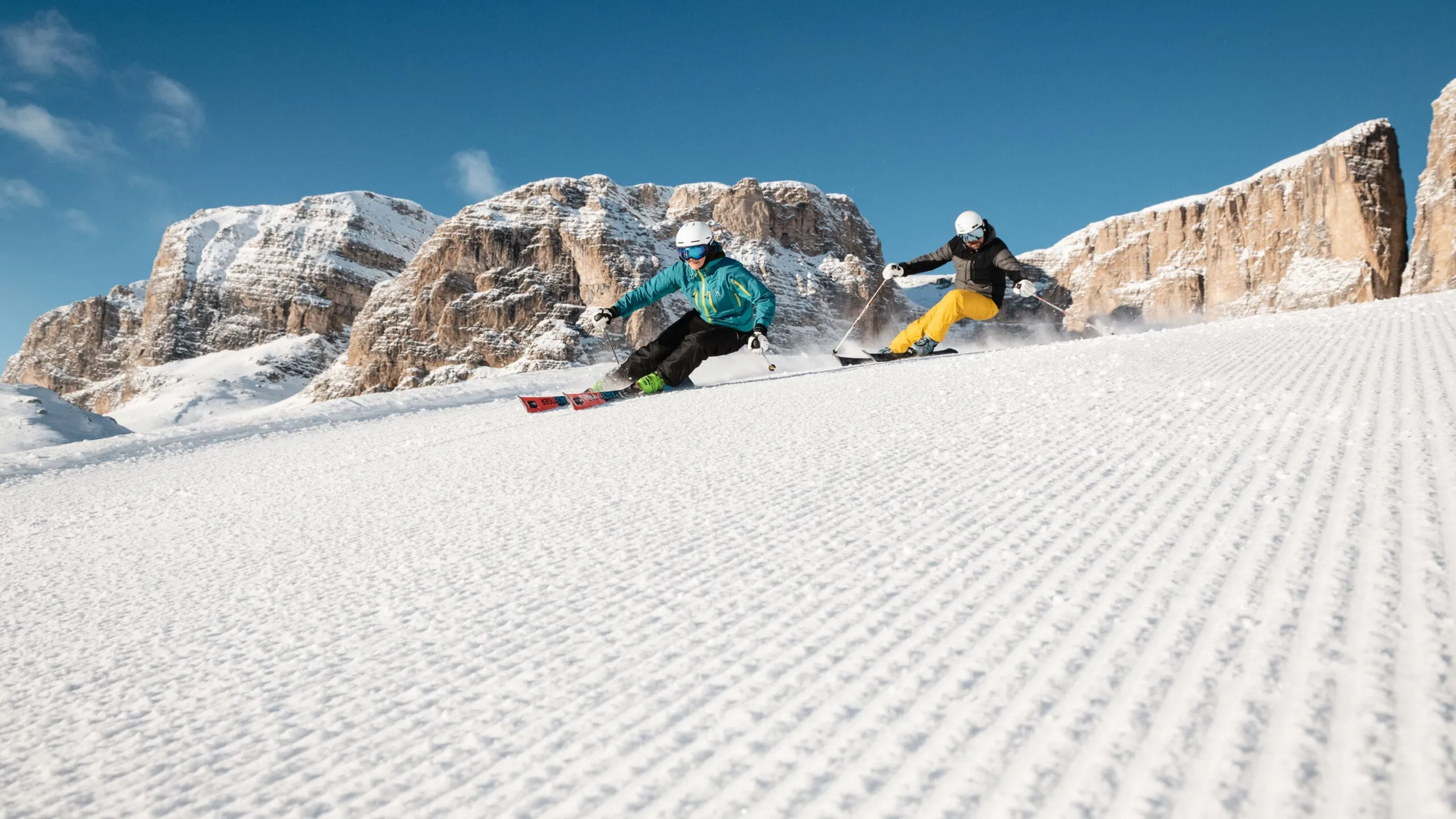 Two skiers carving a snowy slope with rocky cliffs in the background under a clear blue sky.