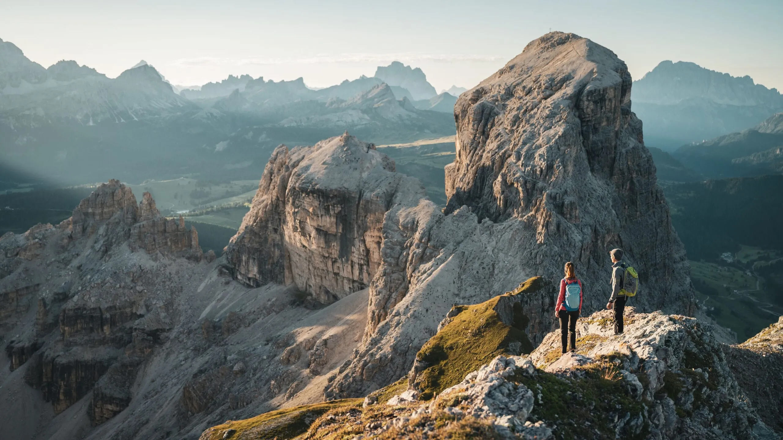 Two hikers with backpacks stand on a rocky mountain ledge, overlooking rugged peaks and a valley below.