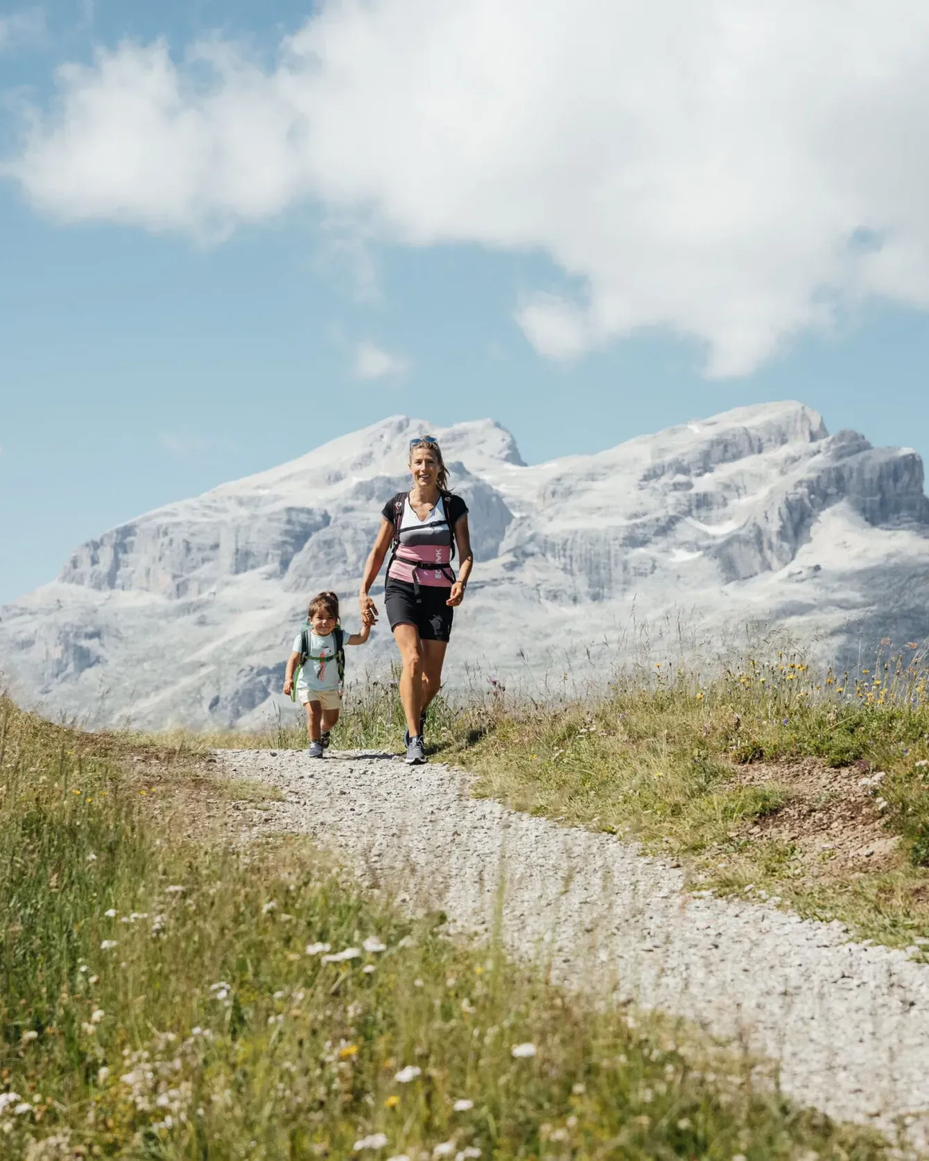Hiking two people, an adult and a child, along a grassy trail with snowy mountains behind.