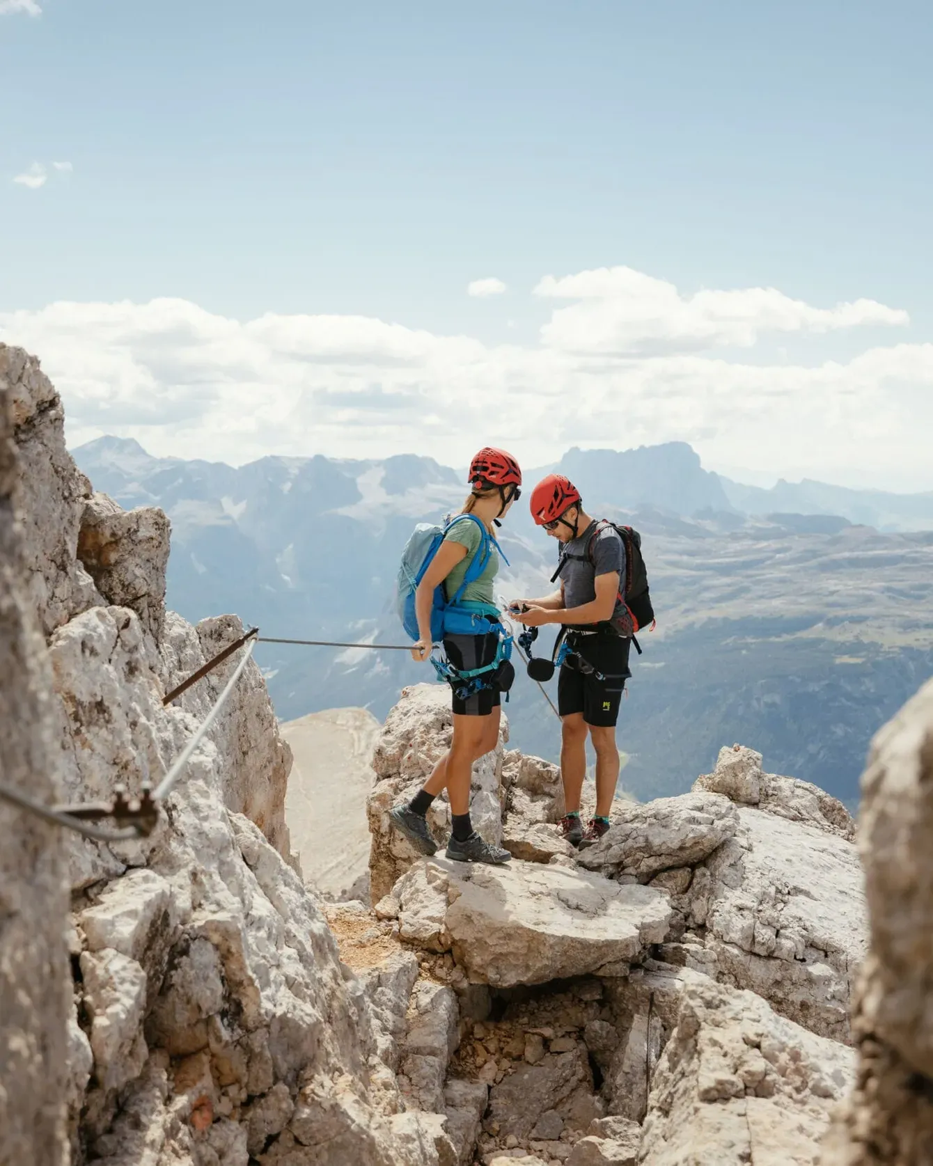 Two climbers with helmets stand on a rocky mountain ridge, checking gear with sweeping alpine valley below.