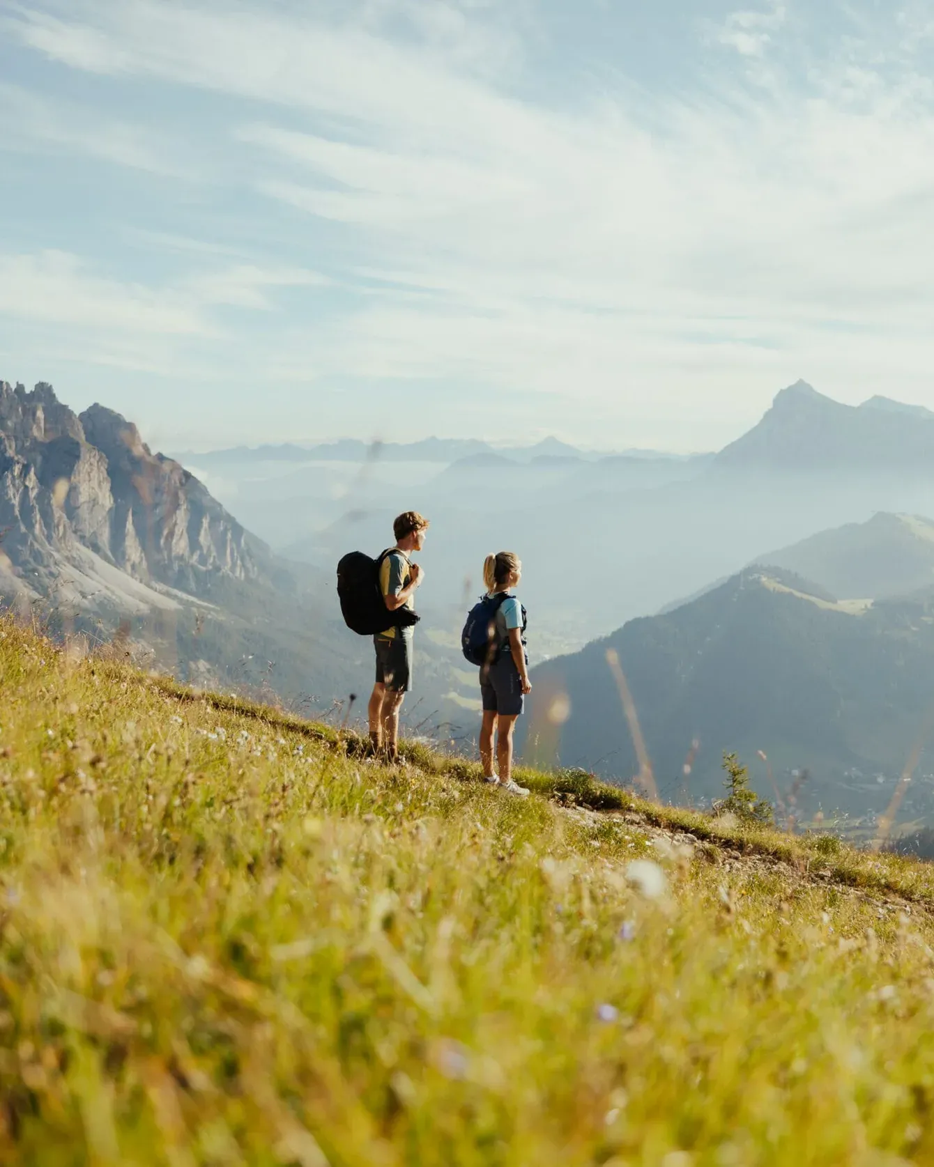 Two hikers with backpacks stand on a grassy hillside overlooking a valley and rugged mountains under a blue sky.