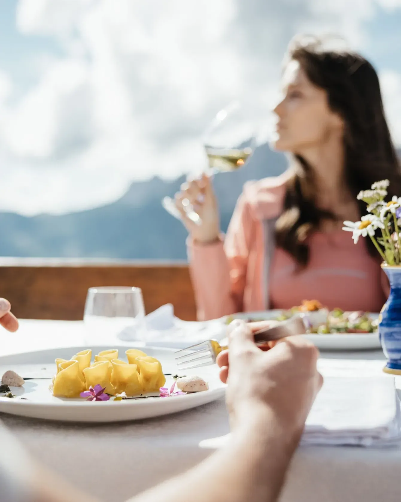 People dining outdoors on a sunny mountain terrace, foreground plate of pasta, wine glass, and flowers in vase.