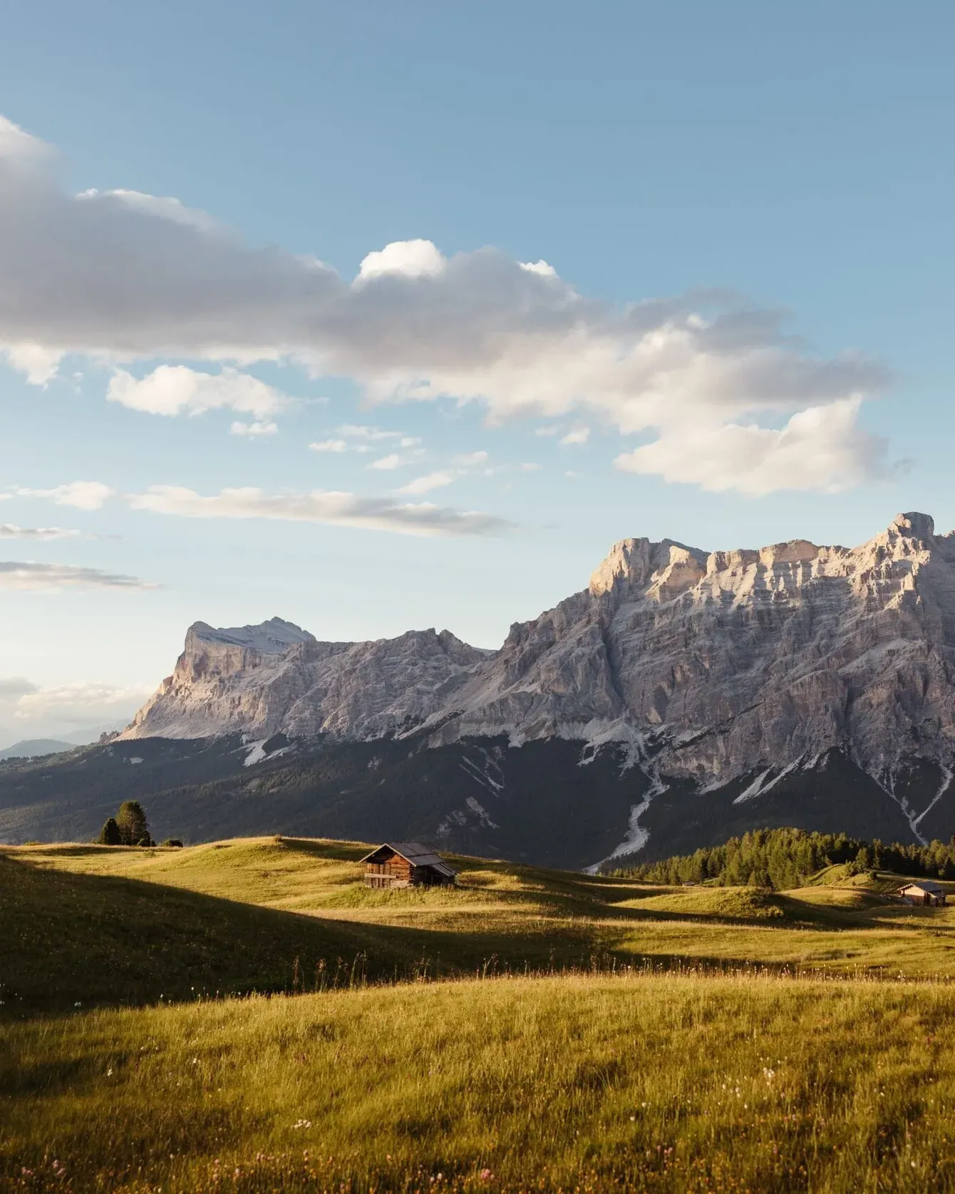 Sunlit grassy meadow with small wooden cabins, vast mountain range in the distance under a blue sky with scattered clouds