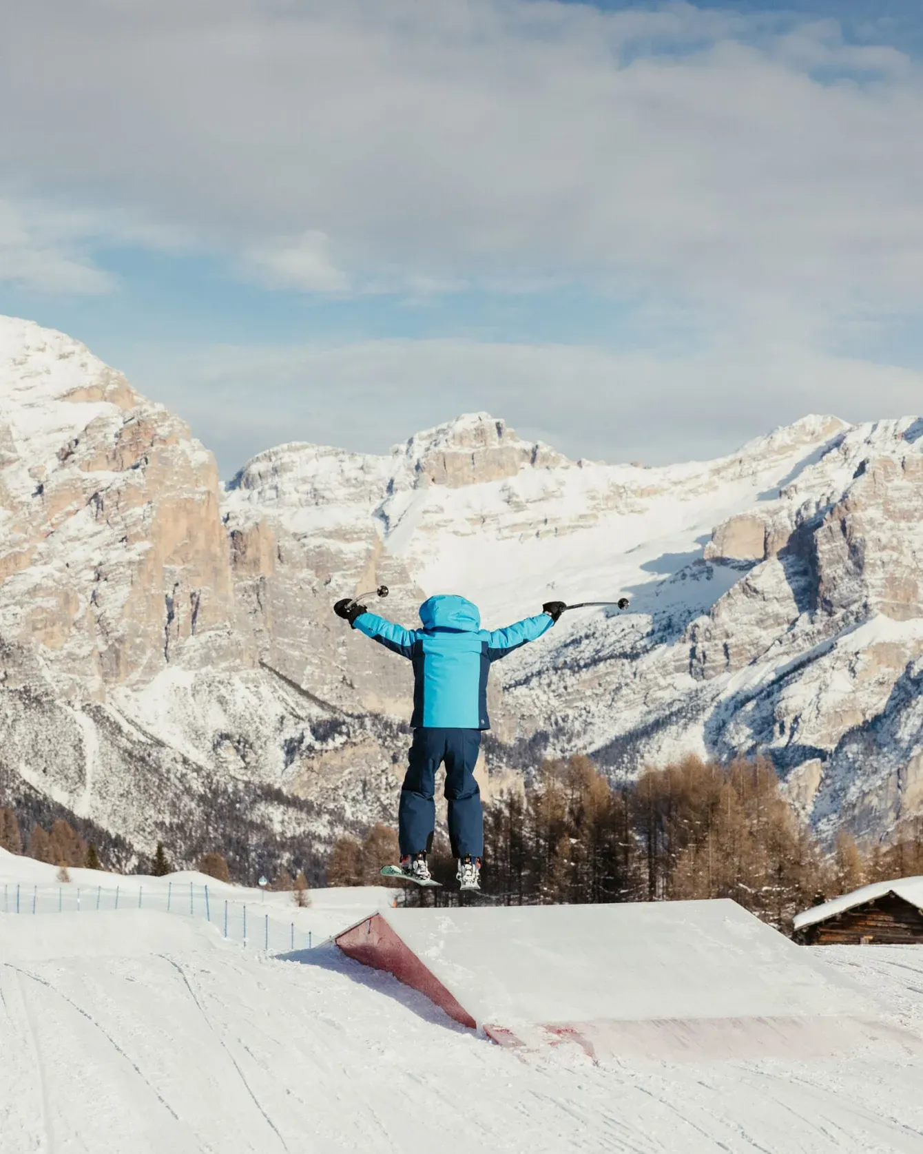 Child in blue jacket jumps with arms spread on a snowy slope against a backdrop of snow-covered mountains.