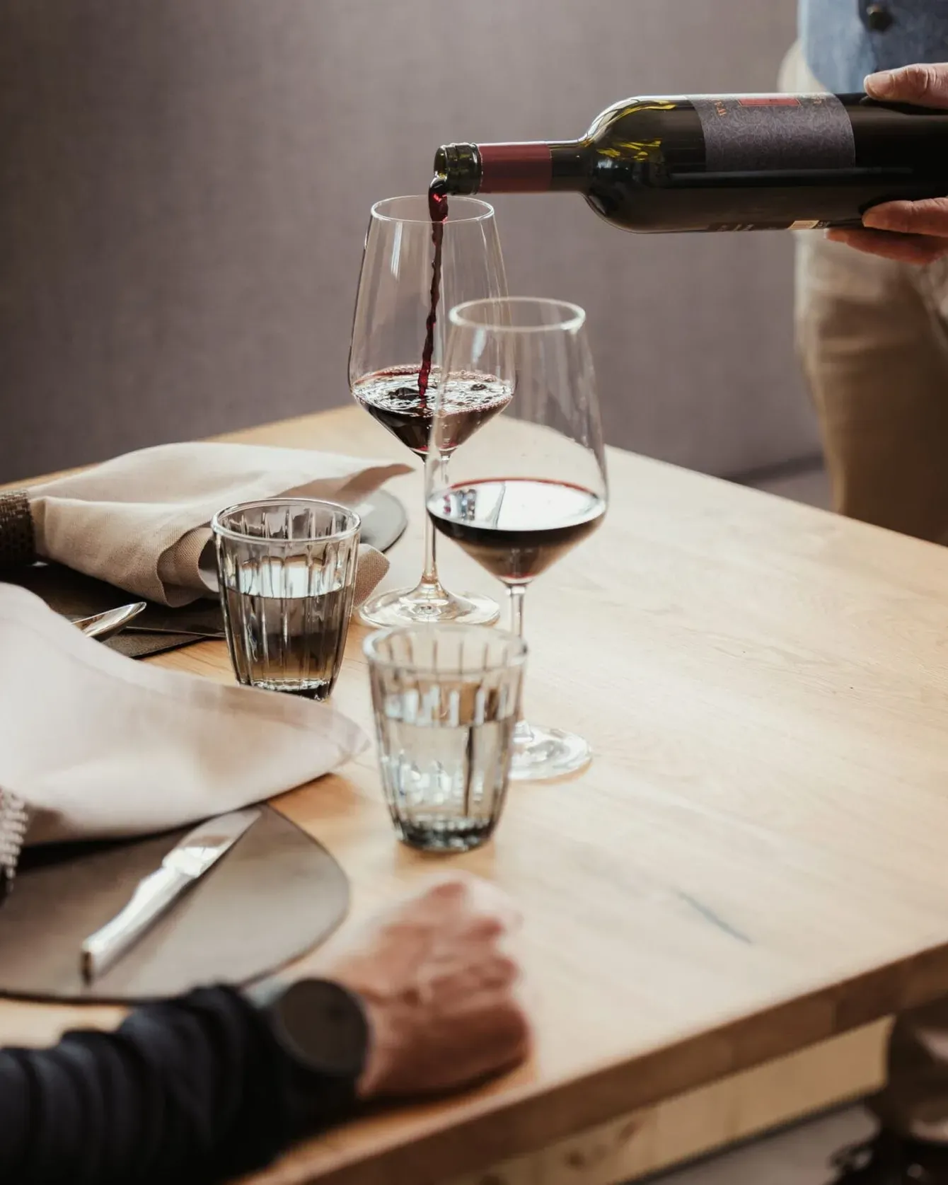 Wine being poured from a bottle into a glass at a wooden dining table with napkins and cutlery.