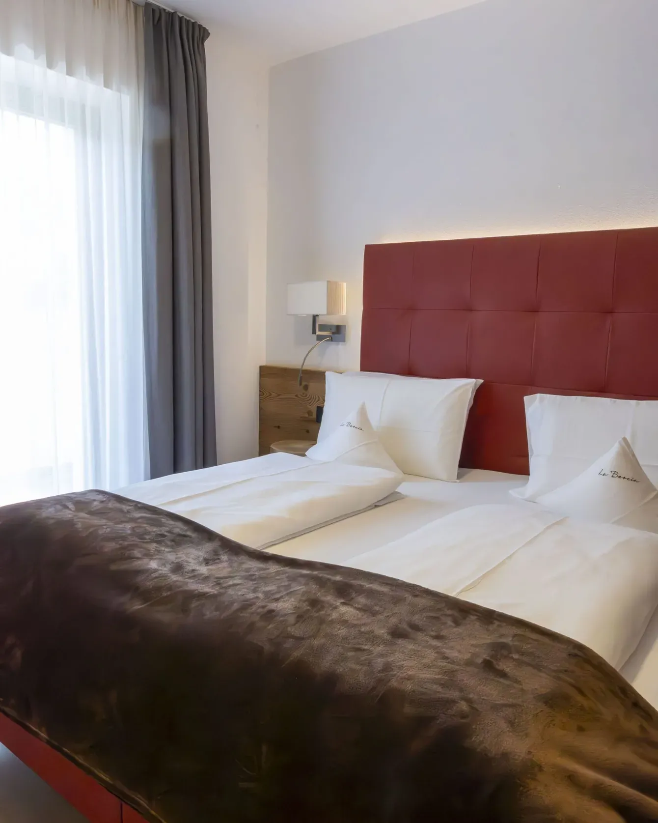 Modern hotel room with a red padded headboard, two white beds, brown silk throw, and sheer curtains by a window.