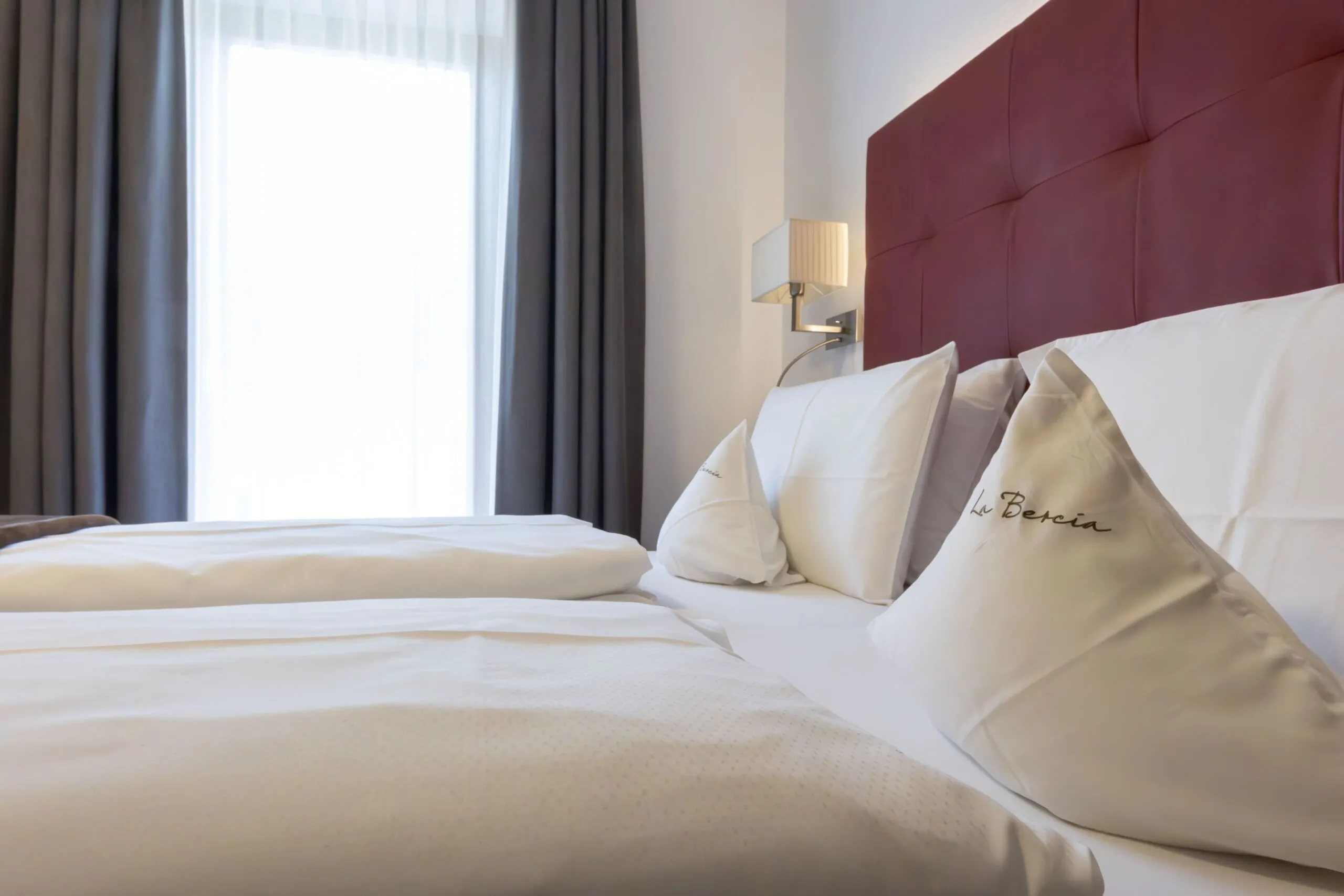 Luxurious hotel bed with white linens, burgundy headboard, and embroidered pillows in a bright room.
