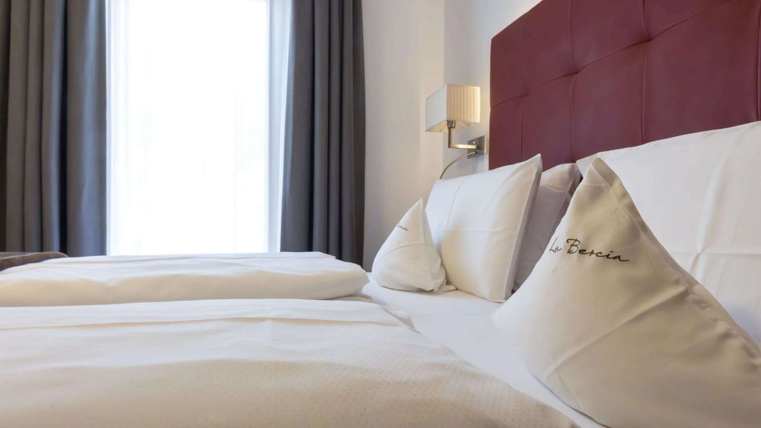 Luxurious hotel bed with white linens, burgundy headboard, and embroidered pillows in a bright room.