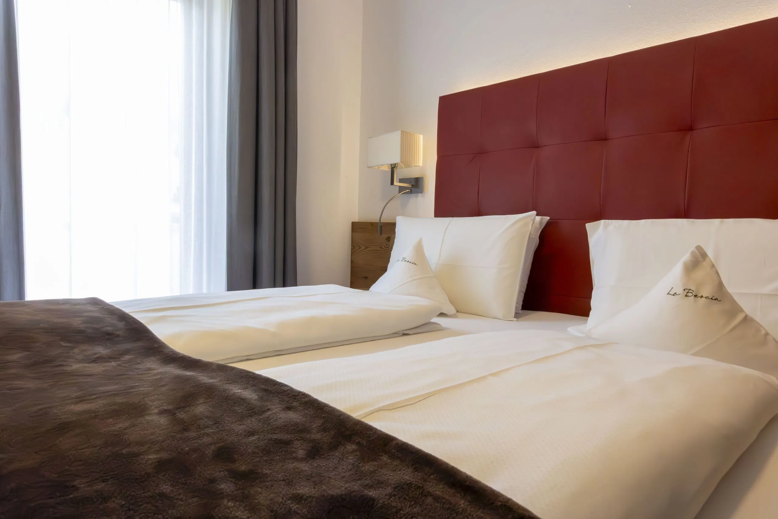 Two neatly made beds with white linens and a deep red tufted headboard in a bright hotel room.