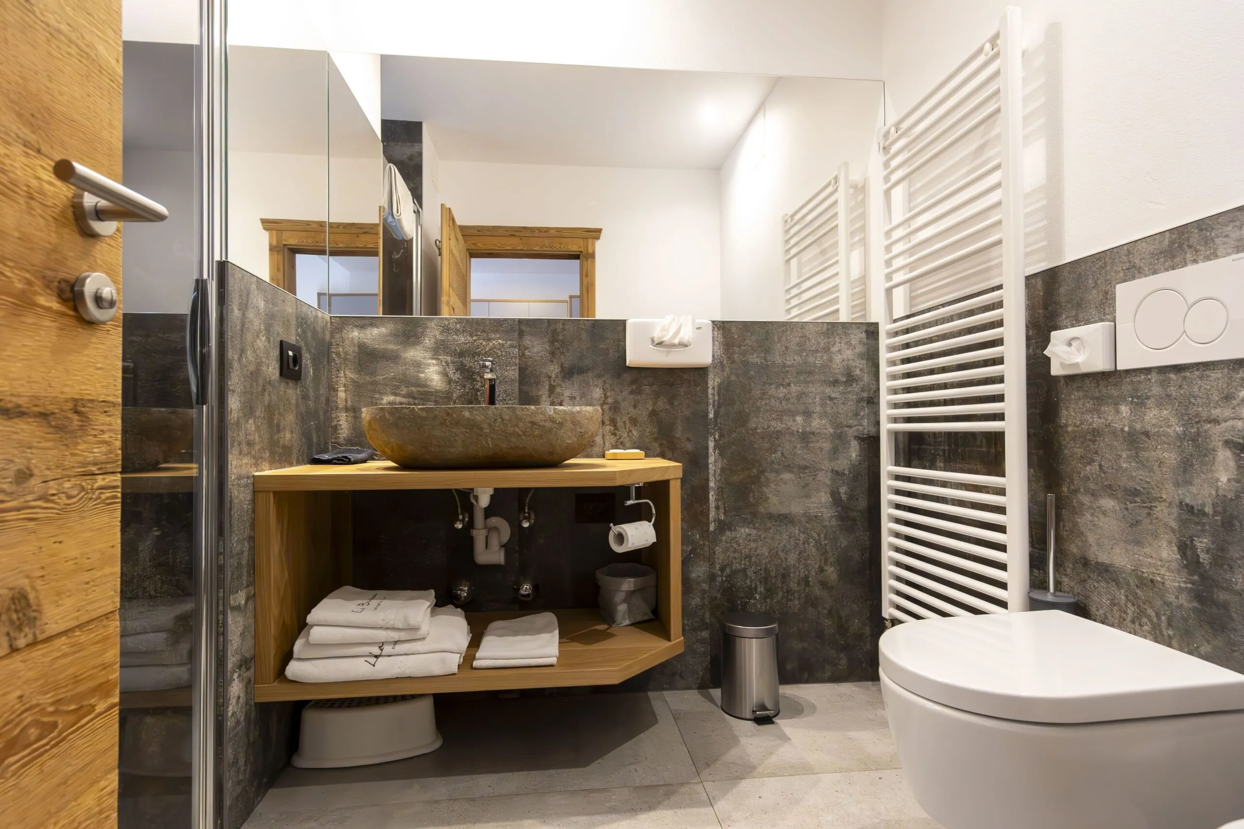 Rustic wooden vanity with stone basin, modern gray tile walls, white toilet, and towel warmer in a bright bathroom.