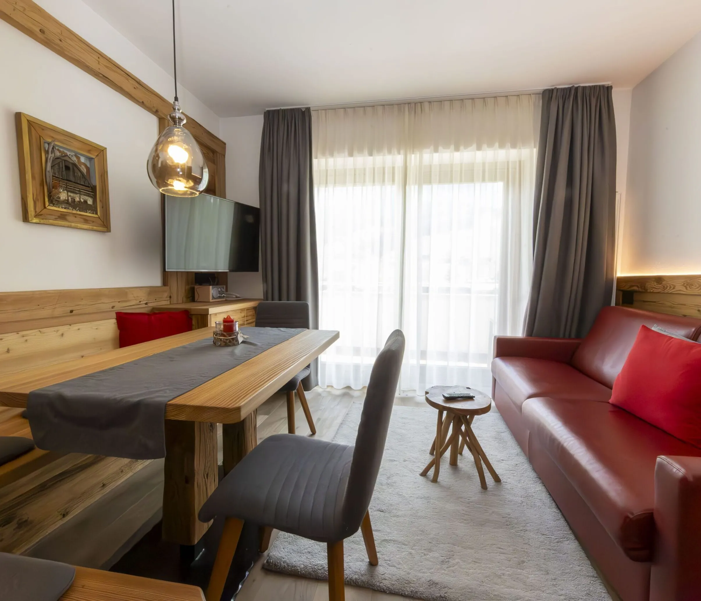 Cozy living area with wooden paneling, red sofa, gray chairs, and large windows with sheer white curtains.