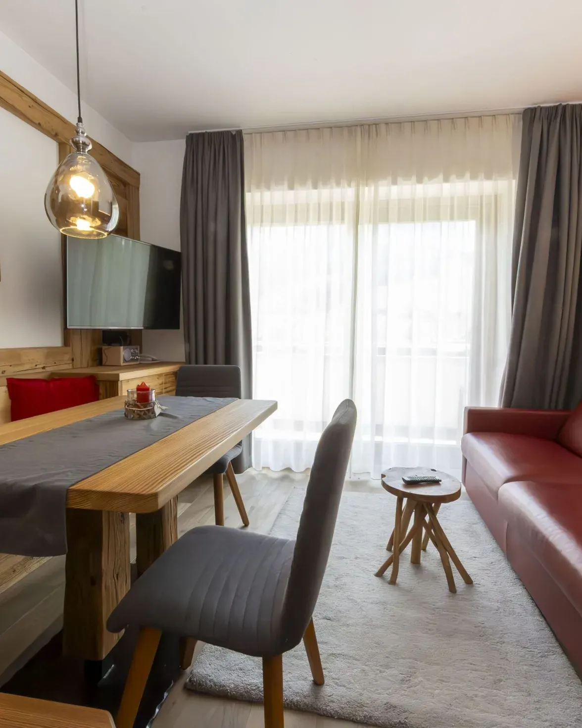 Cozy living area with wooden paneling, red sofa, gray chairs, and large windows with sheer white curtains.