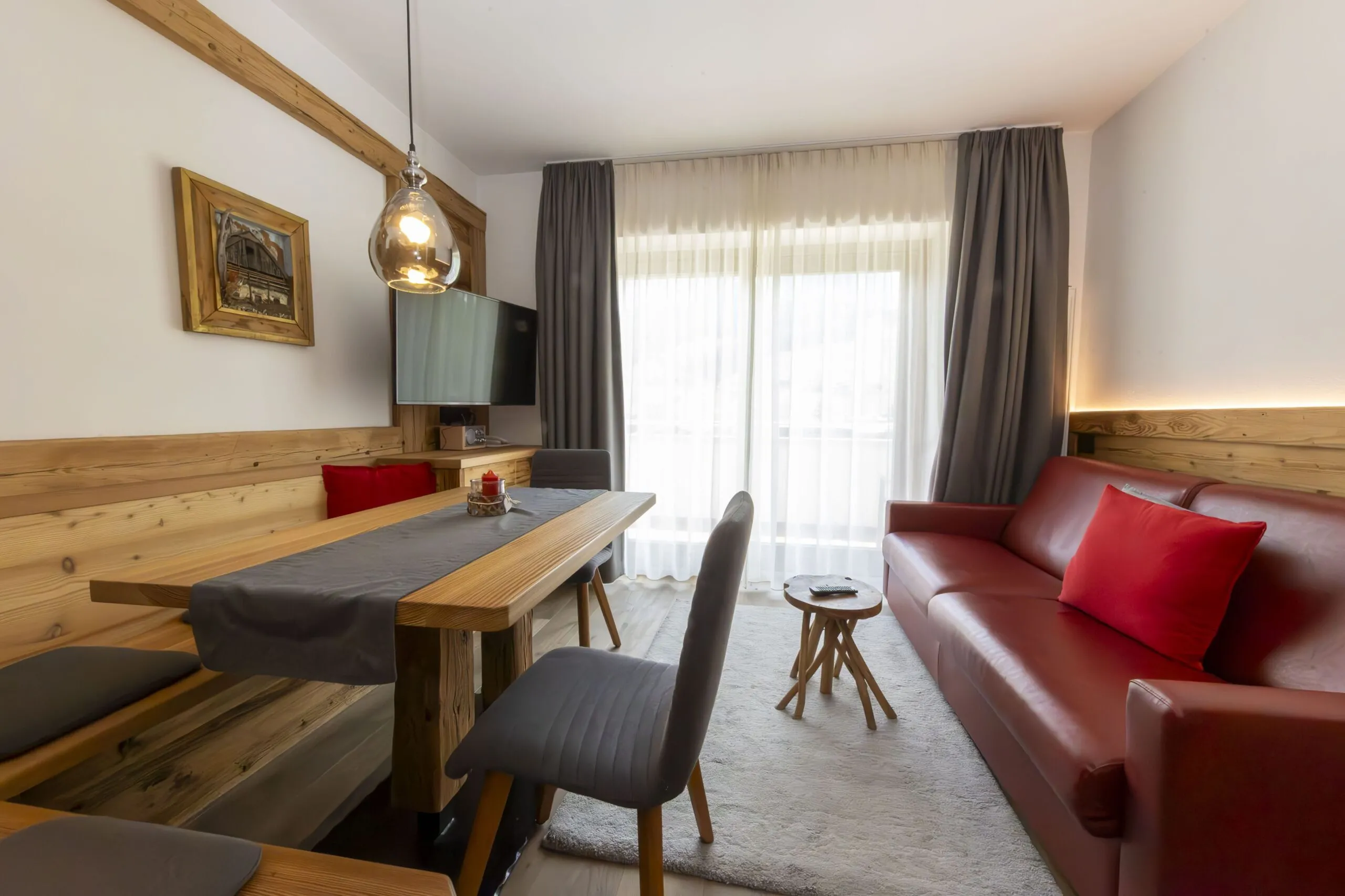 Cozy living area with wooden paneling, red sofa, gray chairs, and large windows with sheer white curtains.