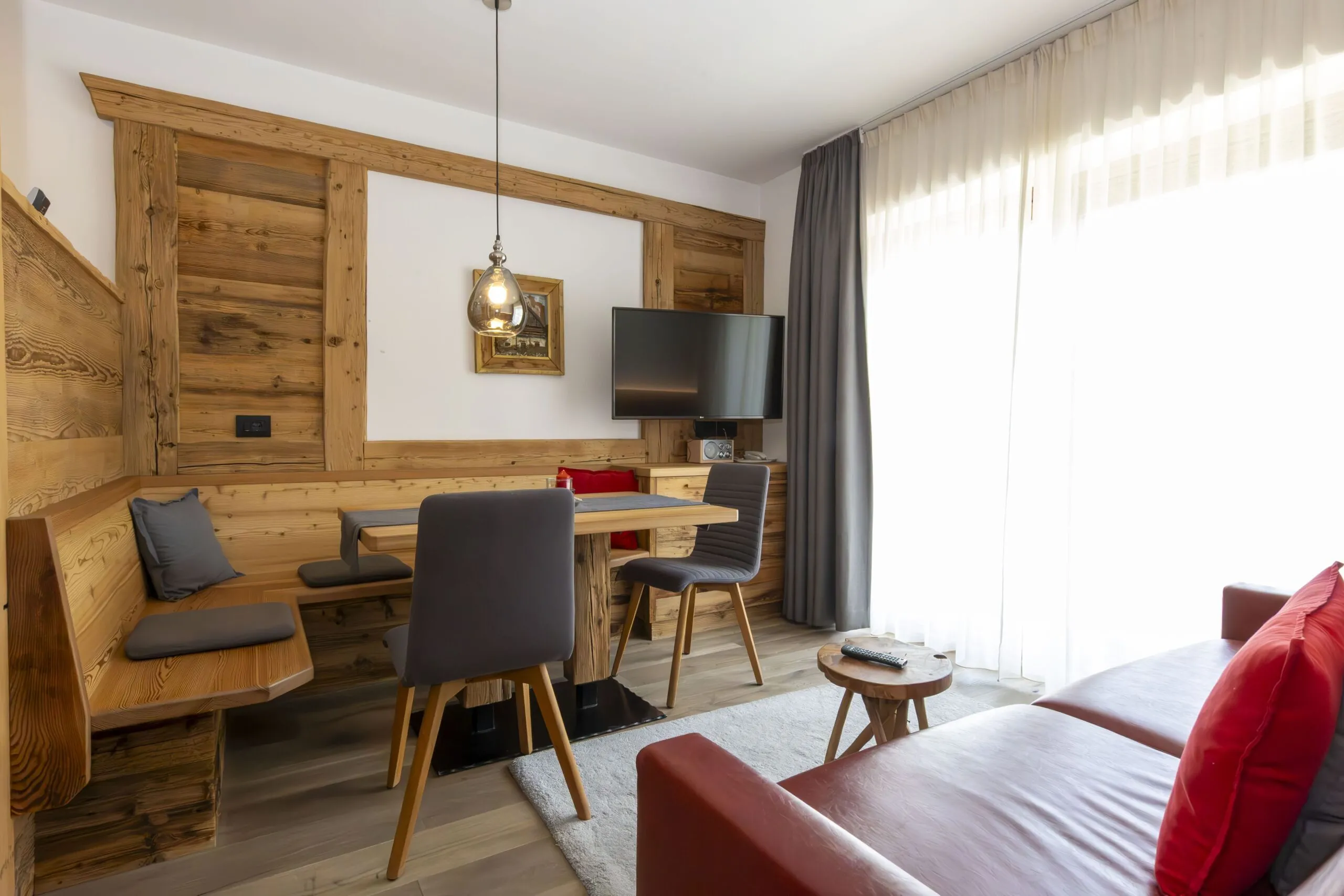 Cozy rustic dining area with wooden paneling, bench seating, gray chairs, wall-mounted TV, and large window with sheer curtains.