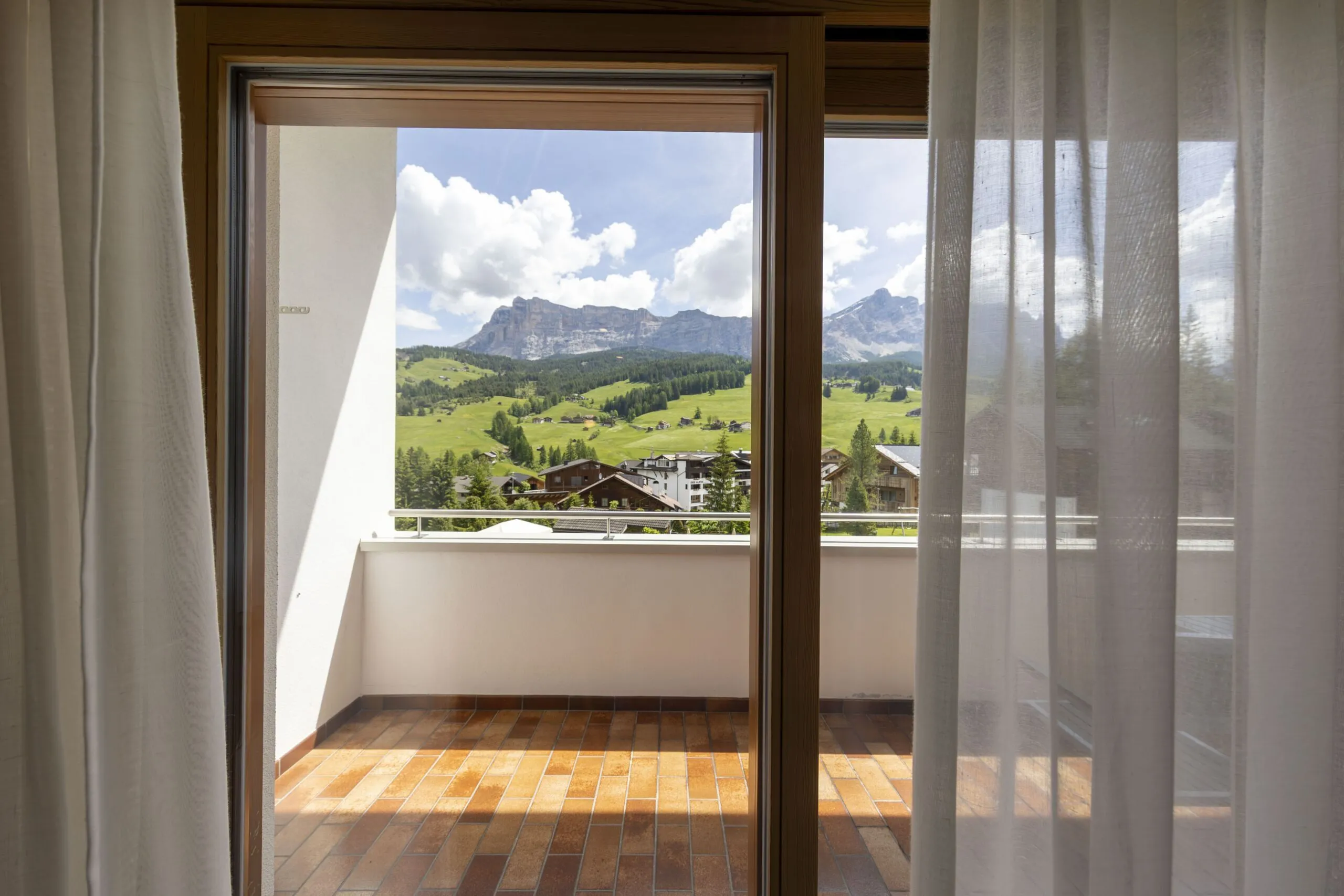 View from a balcony door: green hills, scattered houses, and distant mountains under blue sky.