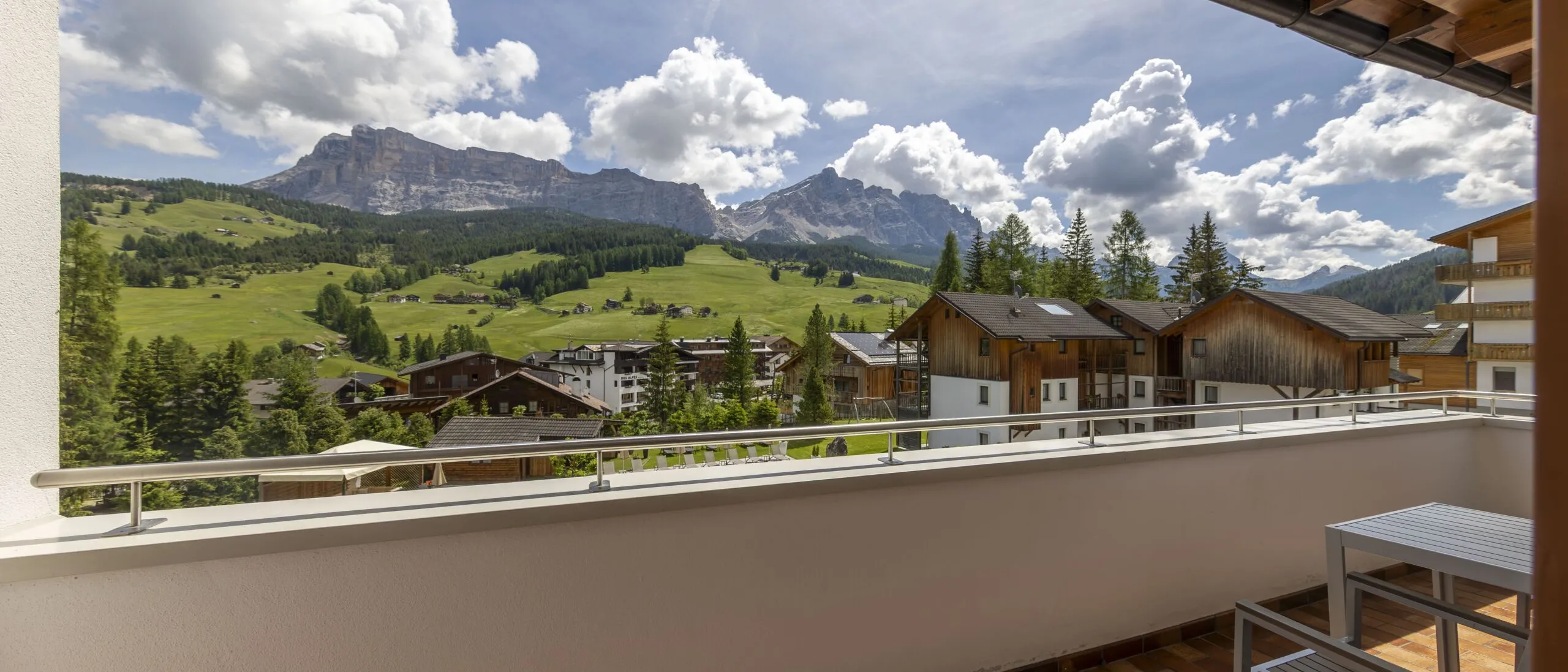 Balcony view of alpine village with wooden houses, green hills, and rugged mountains under a blue sky.