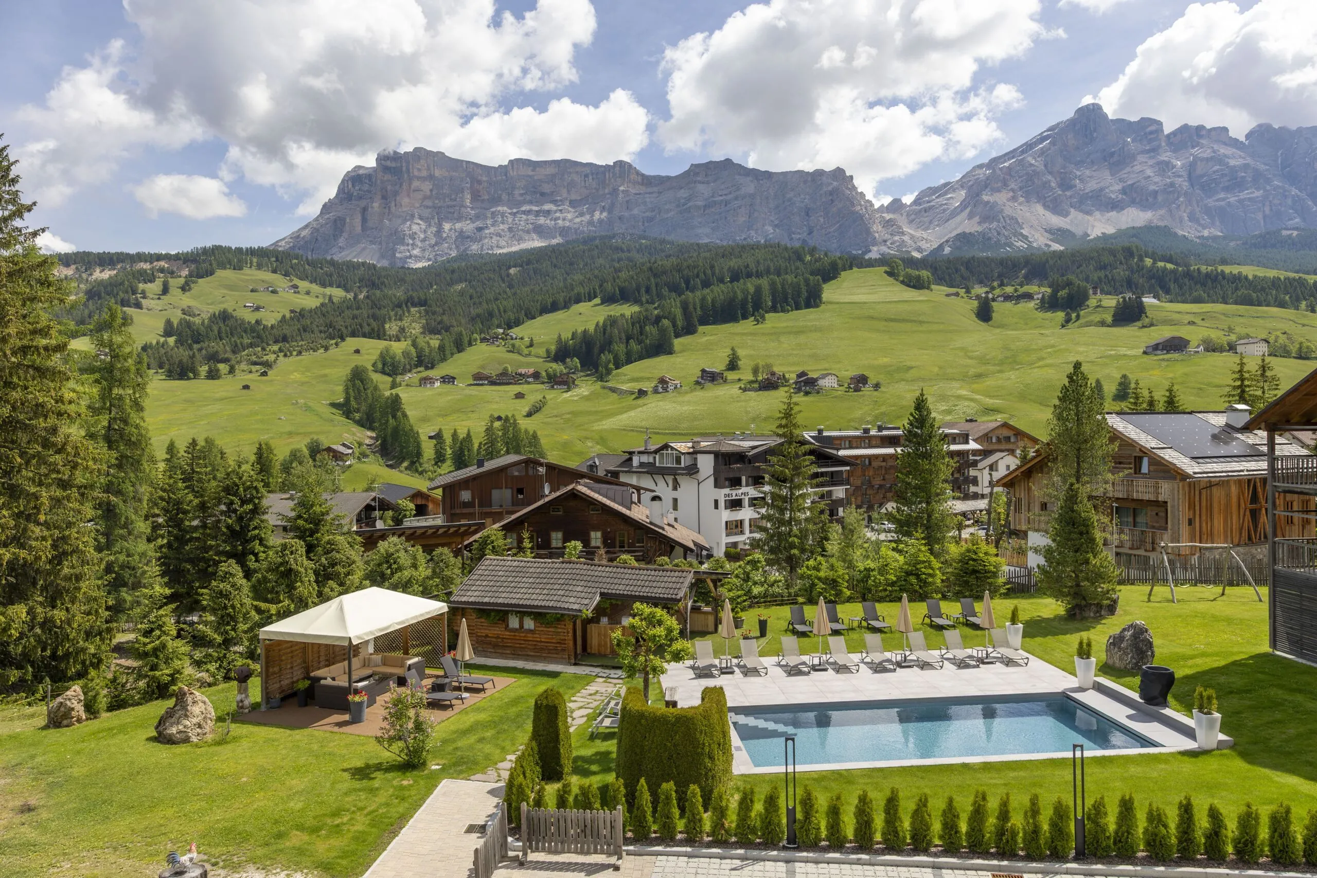 Sunlit alpine village with wooden chalets, green pastures, rows of lounge chairs, and a rectangular pool.
