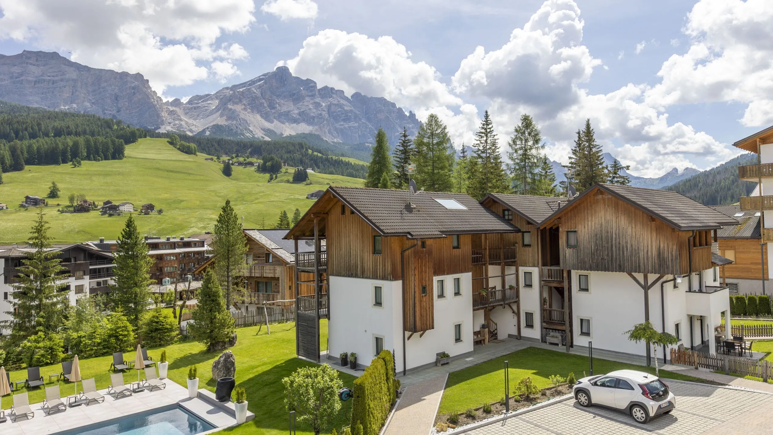 Chalet-style buildings with wooden facades in a mountain village, green hills, and a pool area in the foreground.