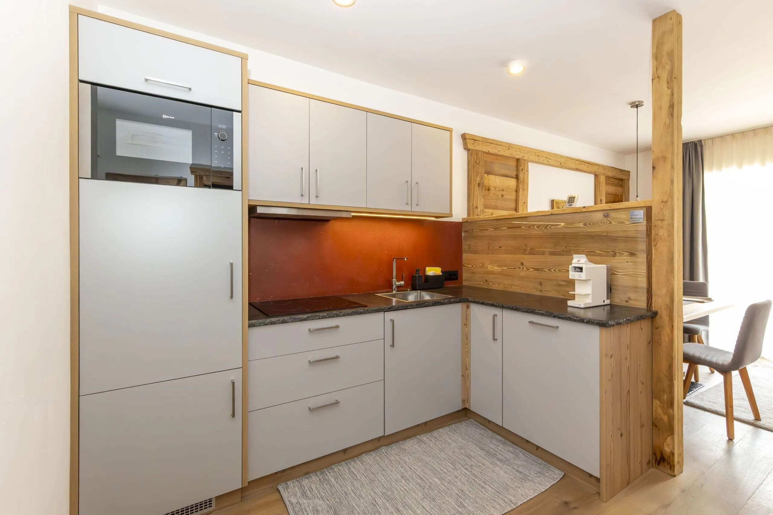 Compact L-shaped kitchen with light gray cabinets, warm wood accents, red backsplash, and small appliance on counter.