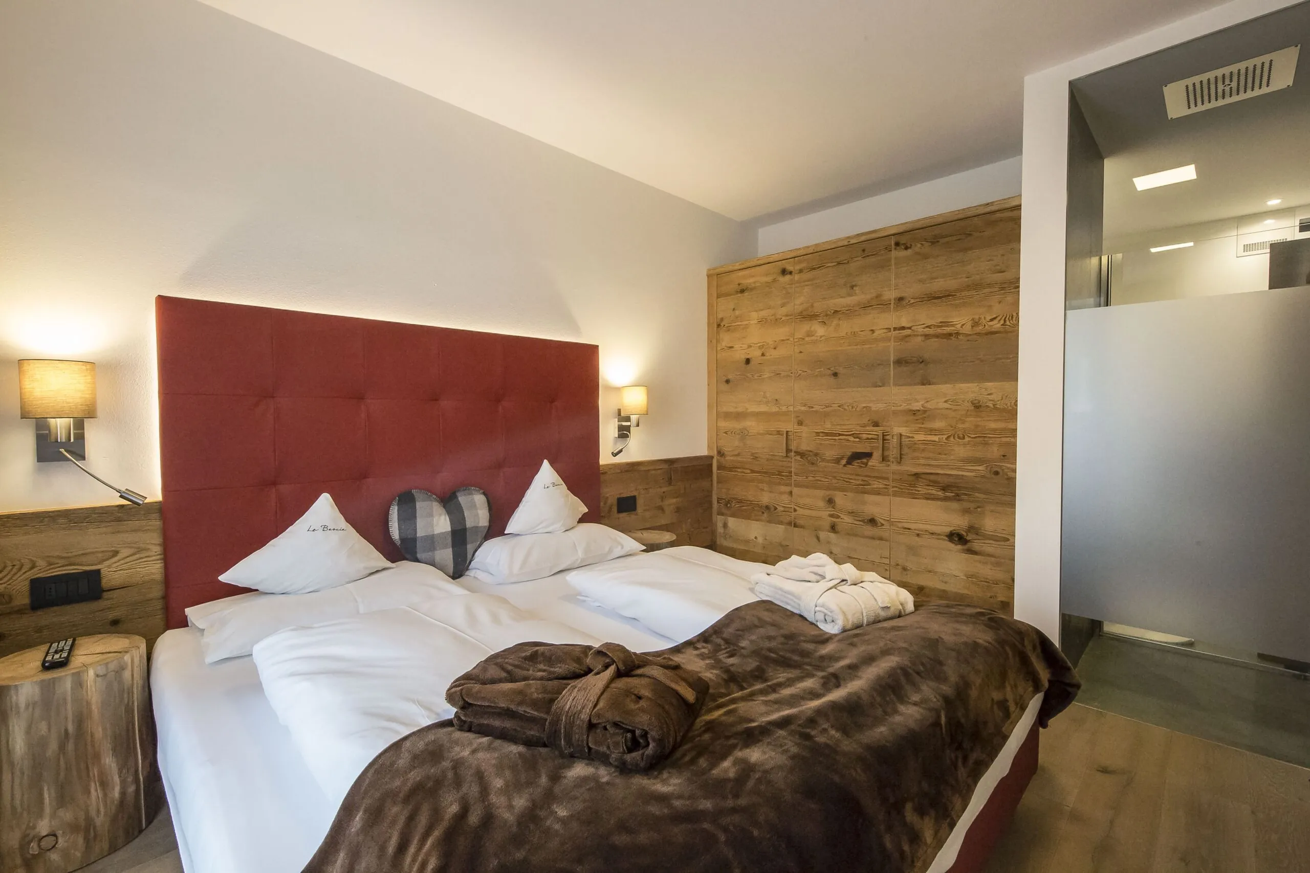 Cozy hotel bedroom with a red padded headboard, two white beds, heart and pillow decor, wooden wardrobe, and soft lighting.